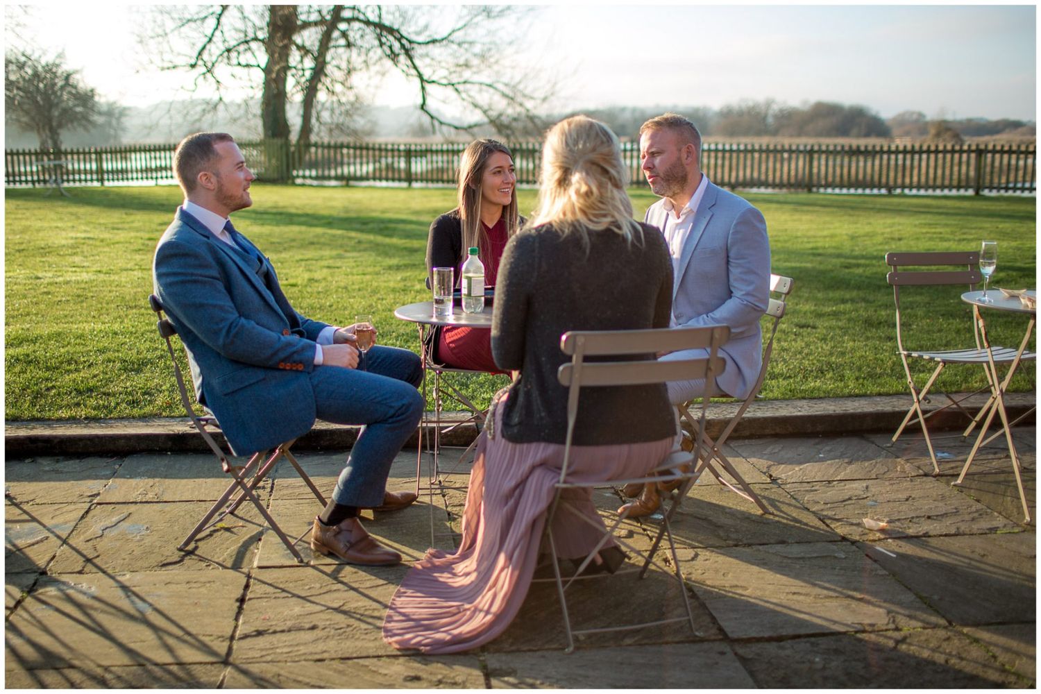 Outdoor wedding gathering with guests seated on chairs in sunny garden setting.