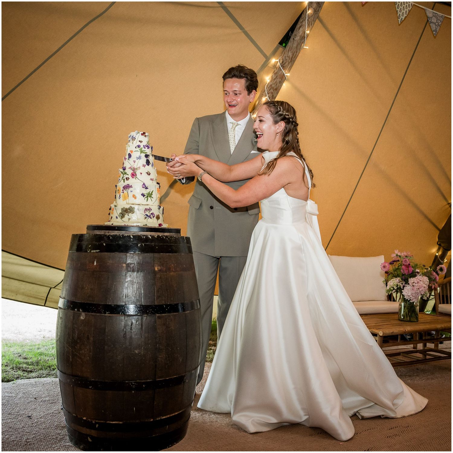 Bride and groom cutting their wedding cake on a wooden barrel inside a tipi tent at the Holywell Estate in Hampshire.
