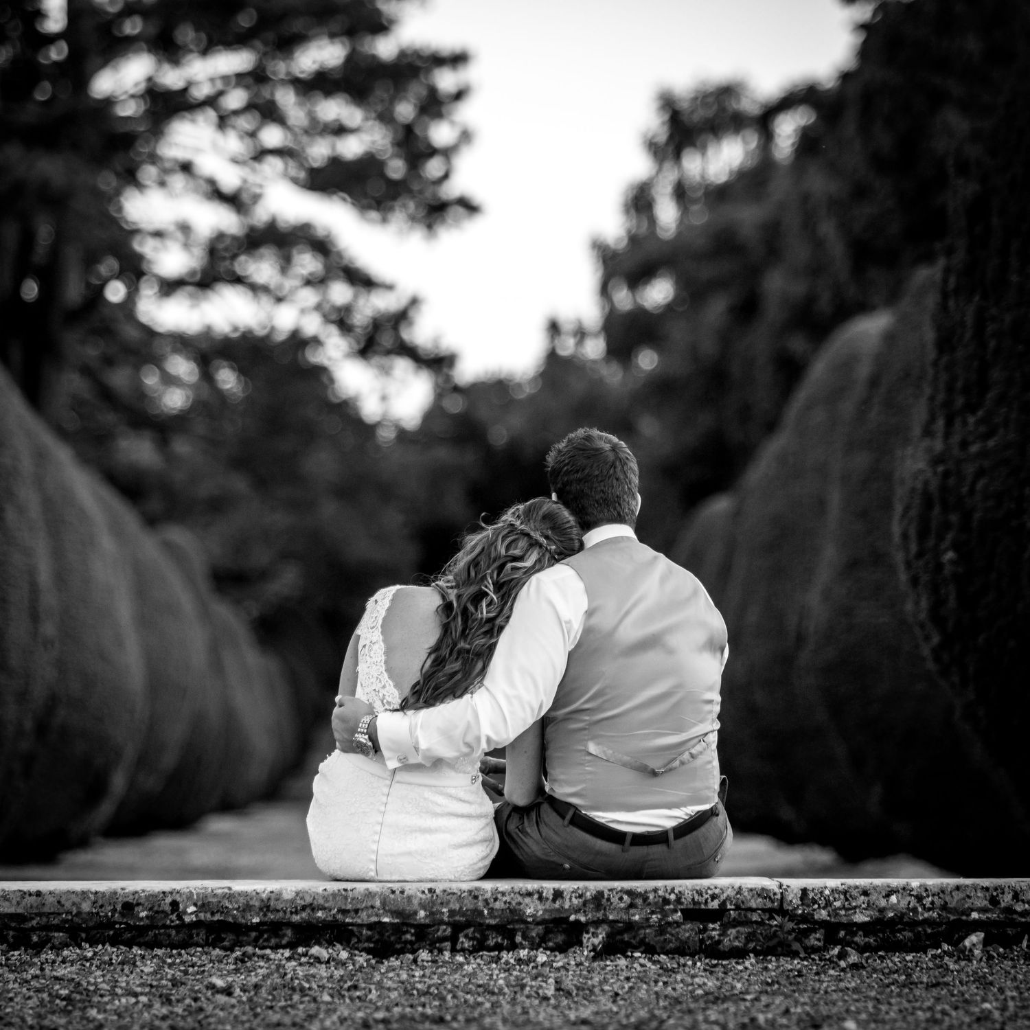 Black and white photo of couple sitting together on stone wall in garden setting.