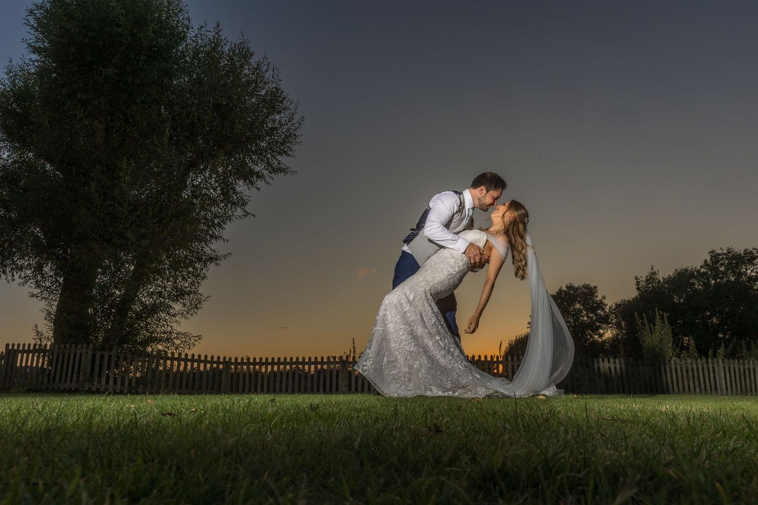 Dramatic sunset wedding portrait with couple in silhouette against fenced garden backdrop.