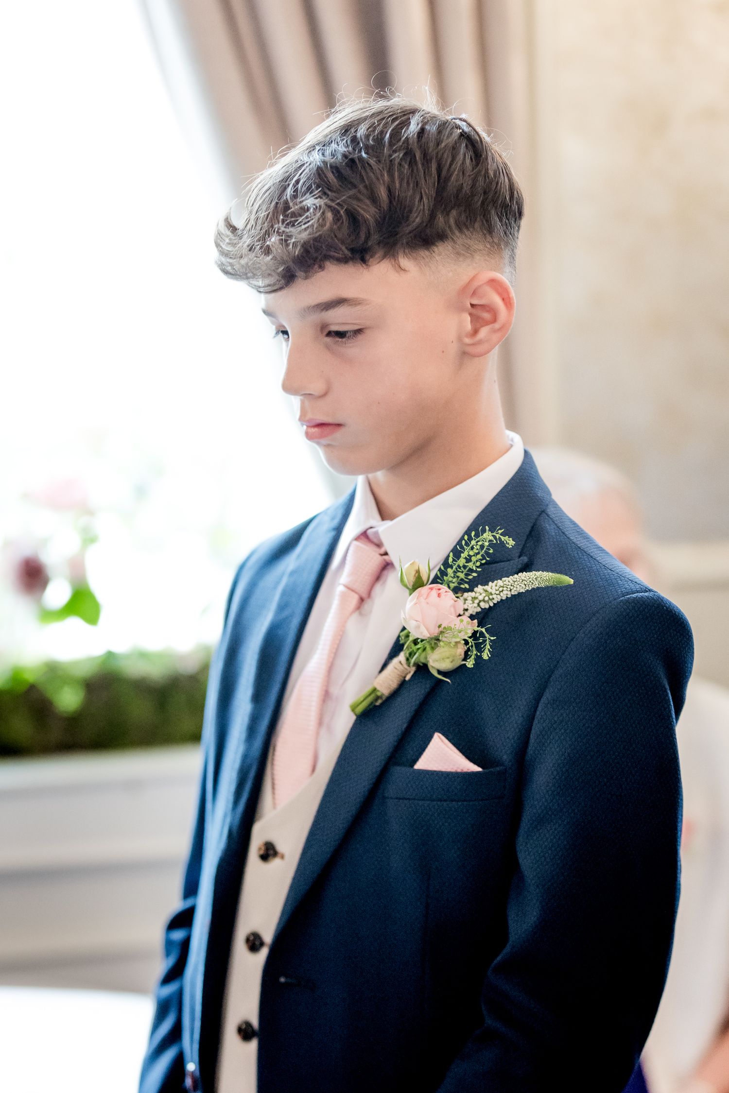 Young groomsman in navy suit with pink tie and boutonniere looking down.