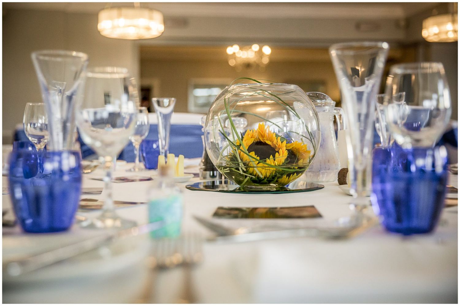 Elegant table setting with blue glassware, sunflower centerpieces, and crystal chandeliers in a banquet hall.