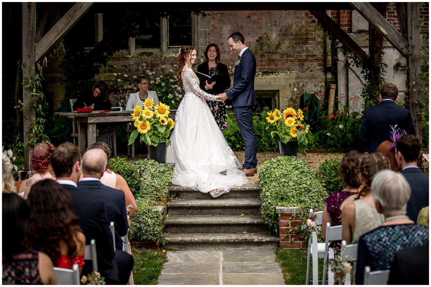 Wedding ceremony guests seated in rows outdoors with sunflowers and garden greenery decorating the venue.
