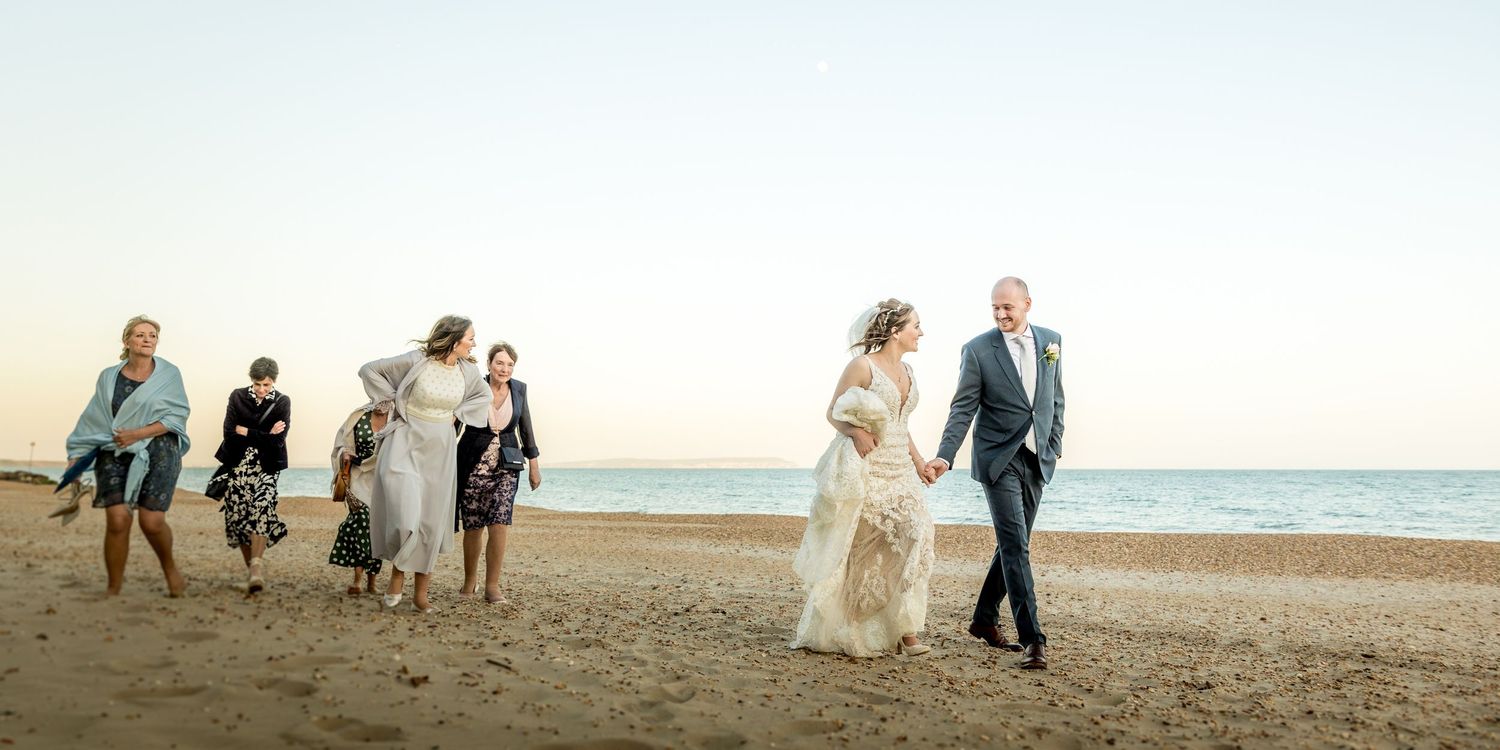 Wedding couple and guests walking along a scenic beach at sunset with ocean waves in the background.