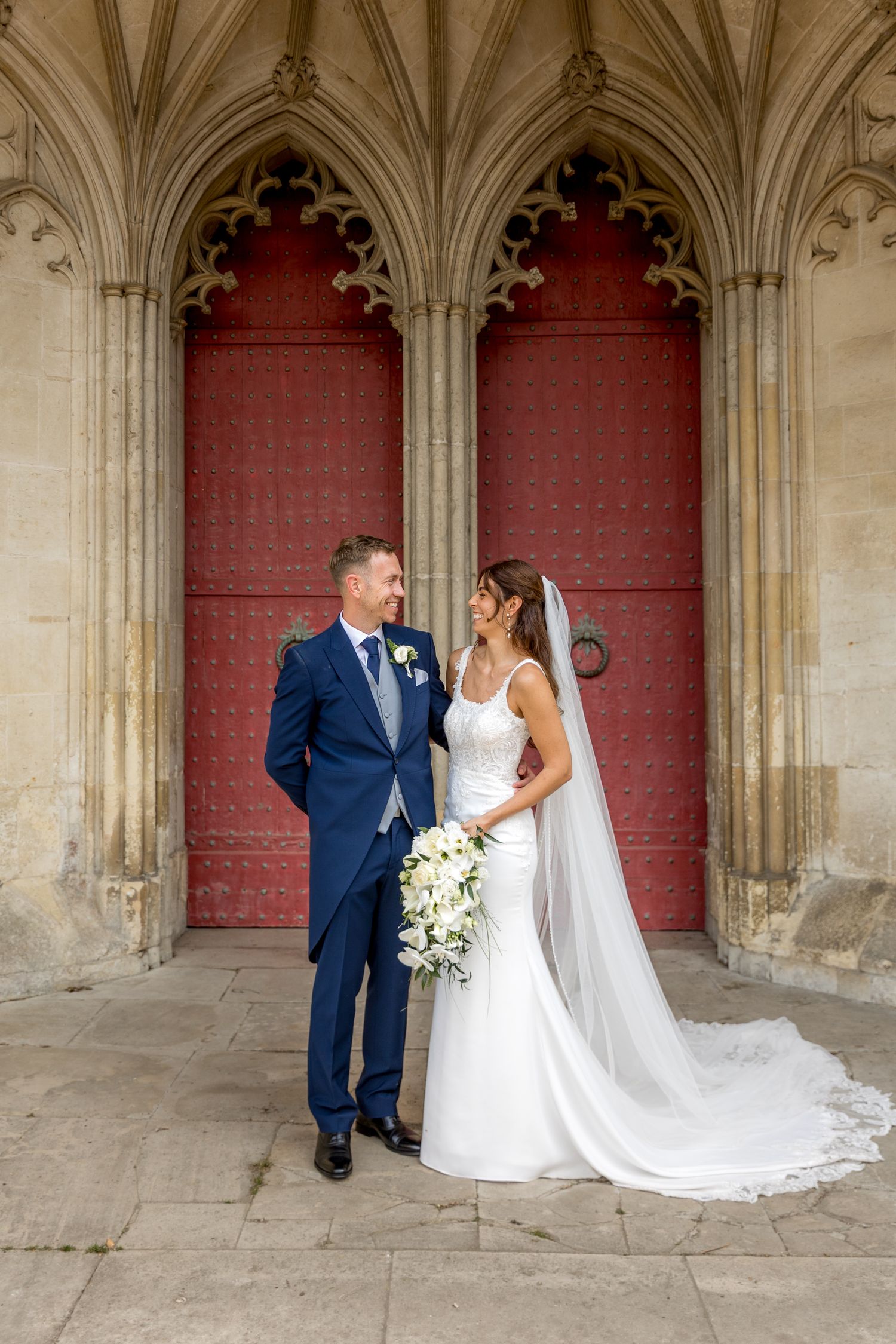 Wedding couple poses under gothic stone archways with red doors and white columns at historic cathedral.