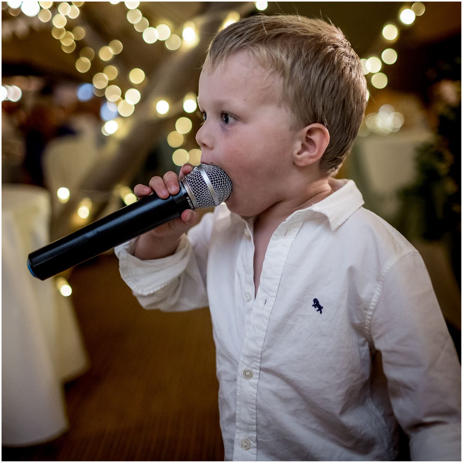 Young child in white shirt sings into microphone at wedding reception with bokeh lights in background.
