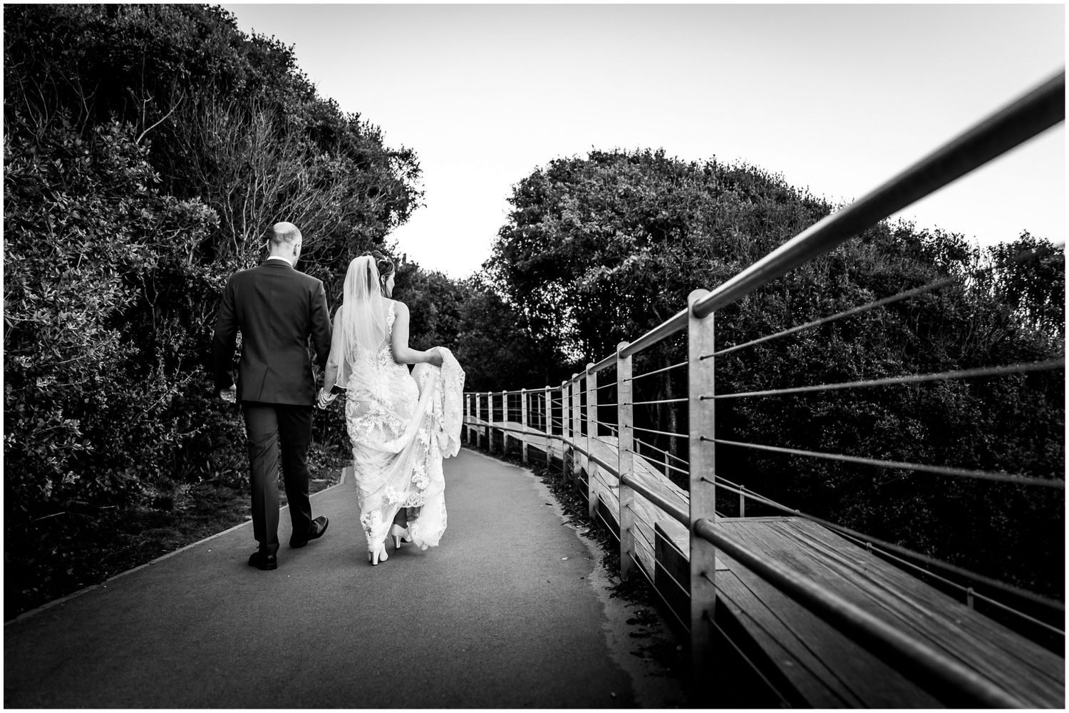 A couple walks along a wooden boardwalk bridge surrounded by lush greenery on their wedding day.