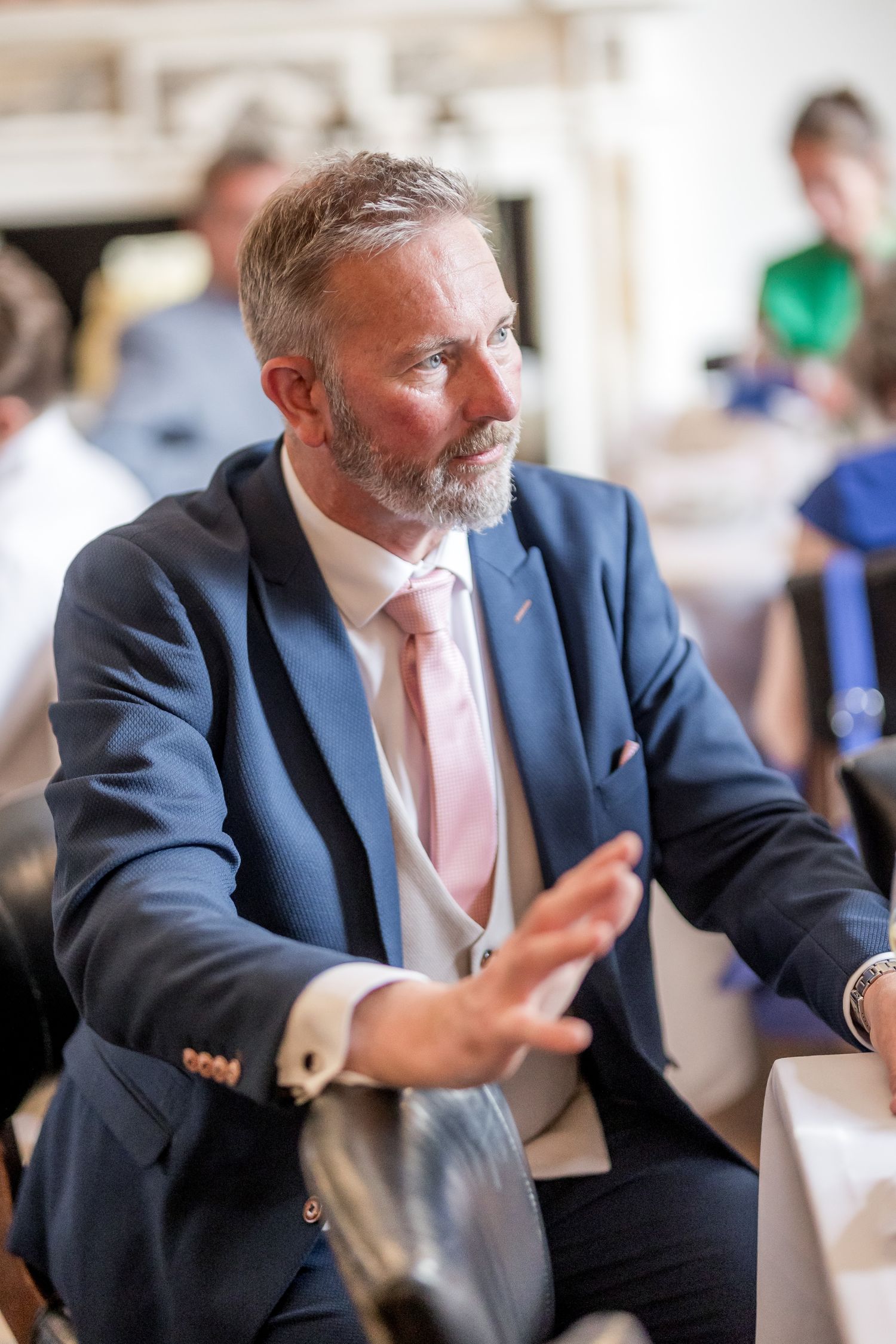Man in navy suit and pink tie gestures animatedly while telling a story.