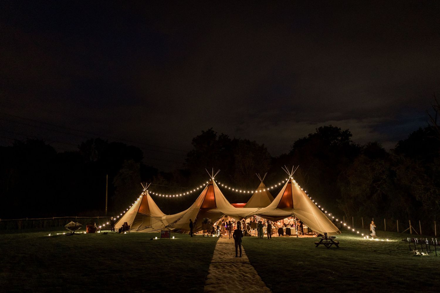Illuminated wedding tent glows warmly against dark night sky with string lights creating magical outdoor atmosphere.