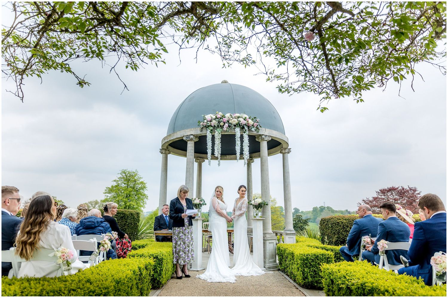 A white wedding gazebo surrounded by lush greenery and guests during an outdoor ceremony.