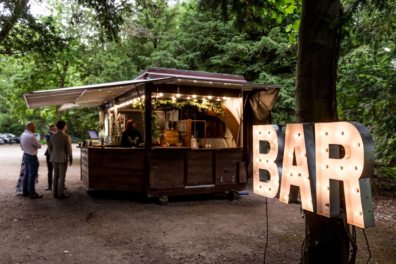 Rustic wooden bar setup with string lights in an outdoor forest venue.