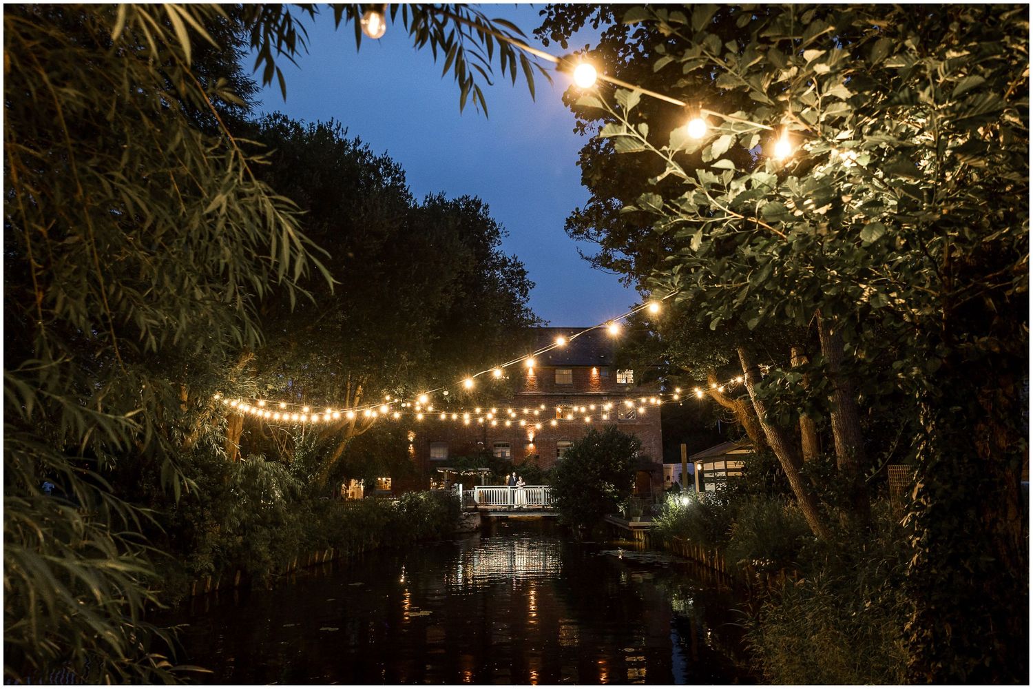 String lights across the river with bride and groom posing on the bridge at Sopley Mill at dusk.
