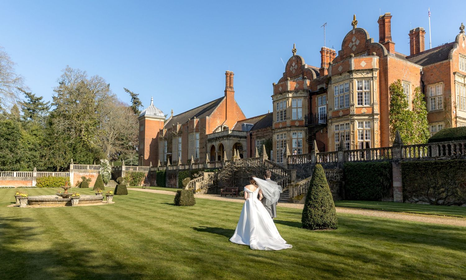 A couple in wedding attire stands on manicured lawn of an elegant Victorian mansion with ornate architecture.