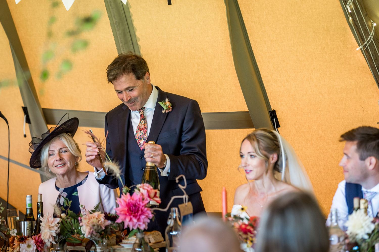 Wedding celebration under a canvas tent with guests enjoying speeches and toasts.