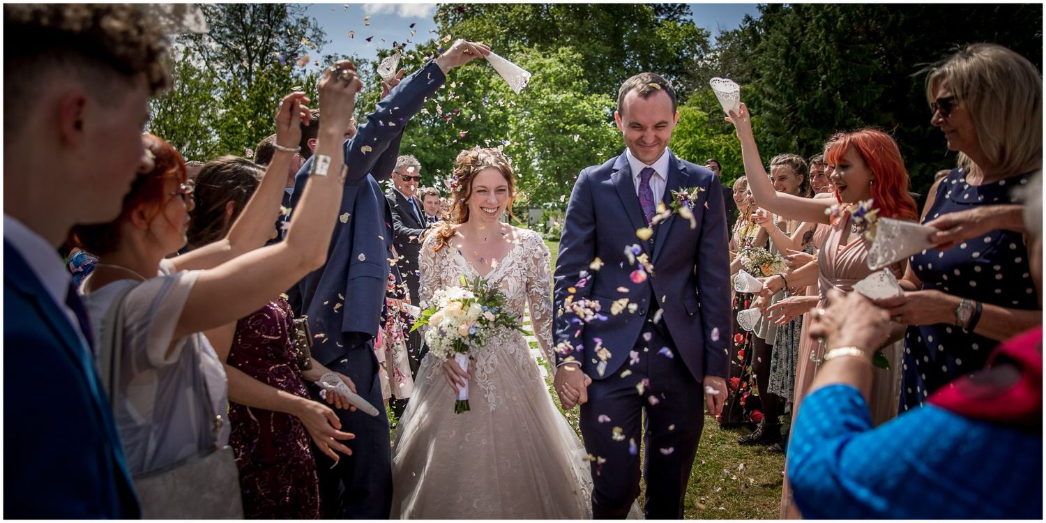 Wedding guests celebrate by throwing confetti and flower petals as newlyweds walk through garden pathway.