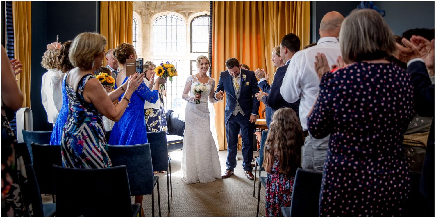 Wedding celebration scene with guests mingling and dancing in a sunlit room with floor-to-ceiling windows.