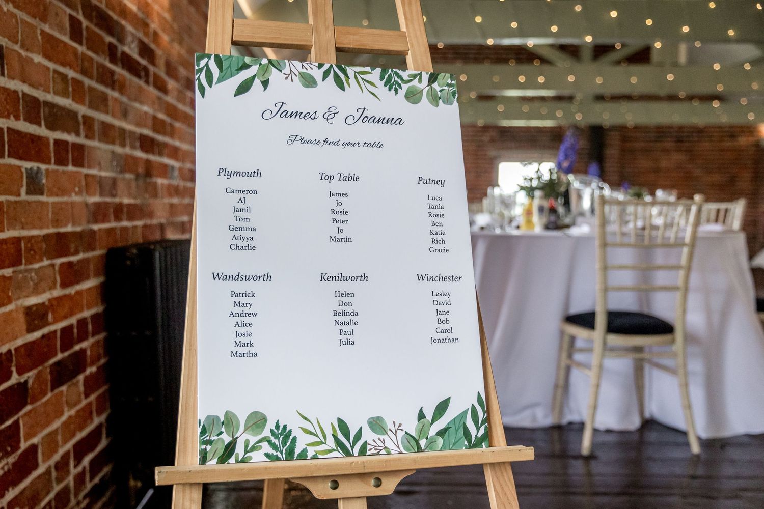 Wedding seating chart display with greenery design on easel in brick venue.