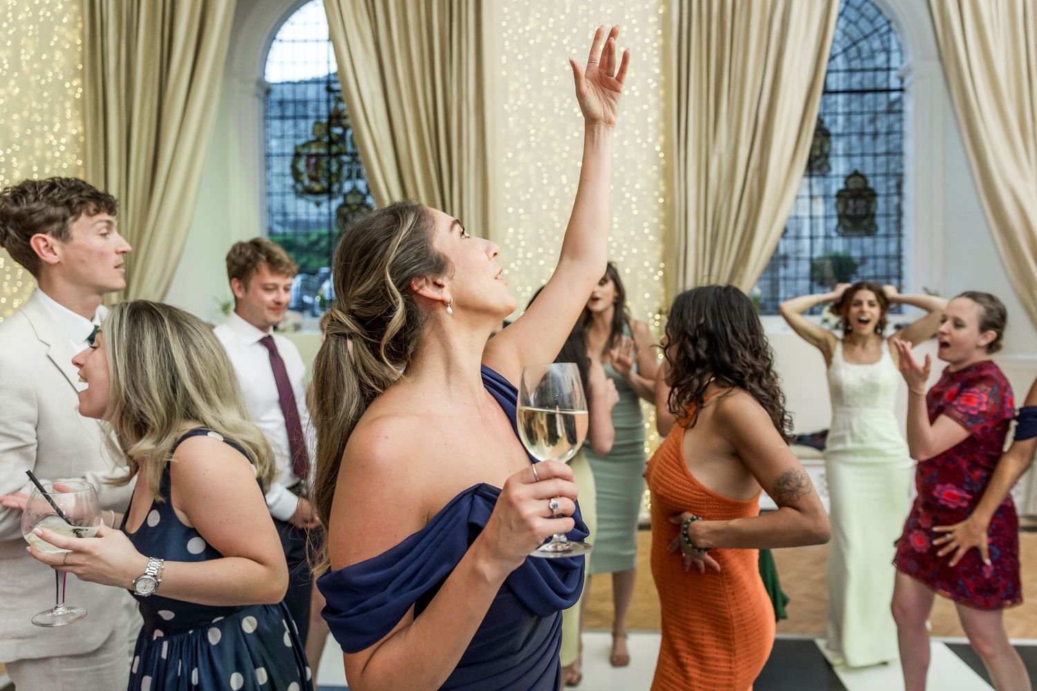 Wedding guests dance energetically in an elegant room with arched windows and cream curtains.
