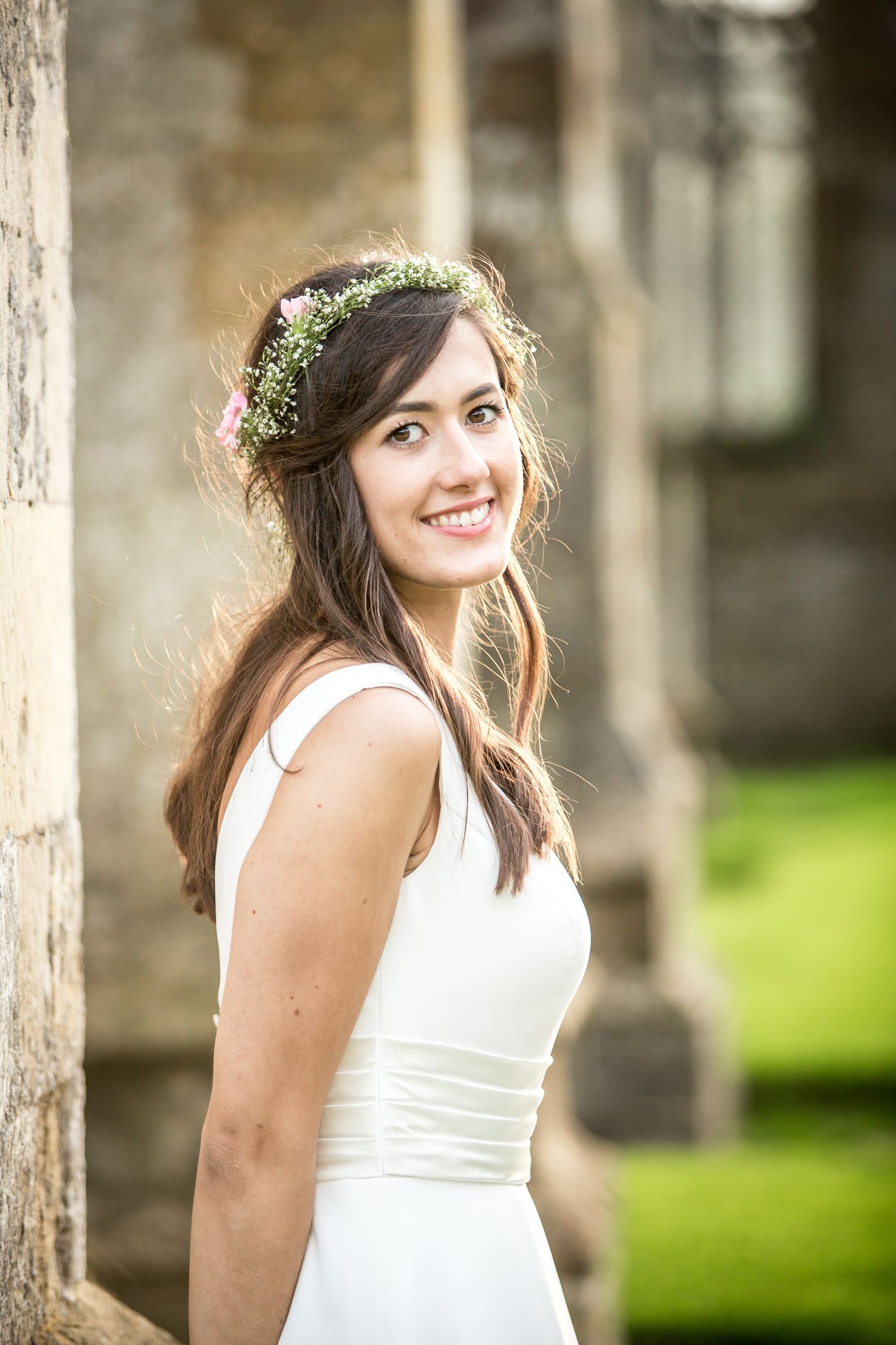A woman in a white dress poses in front of stone columns wearing a floral crown.