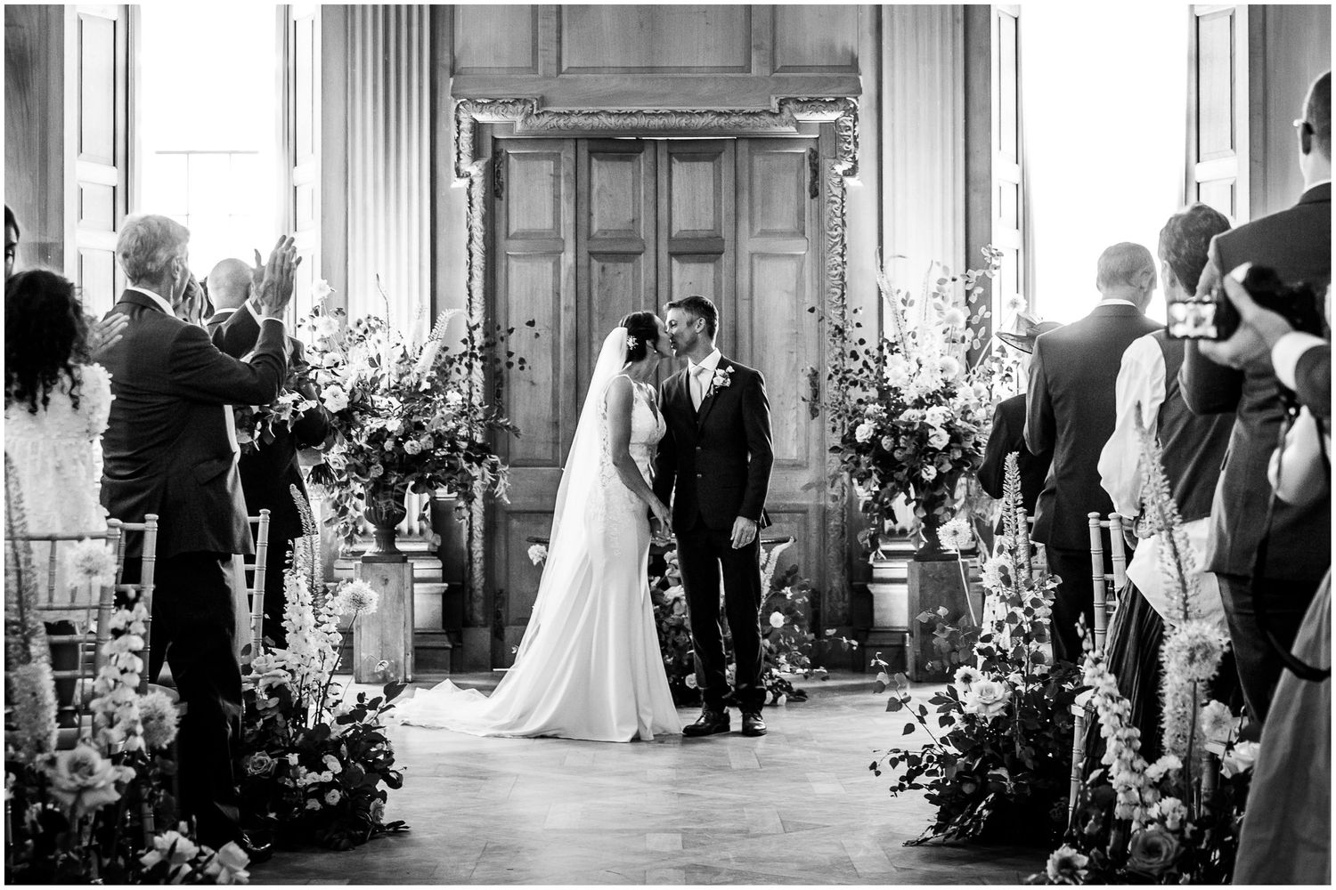 Black and white wedding ceremony photo of bride and groom kissing at altar surrounded by floral arrangements at Walhampton School