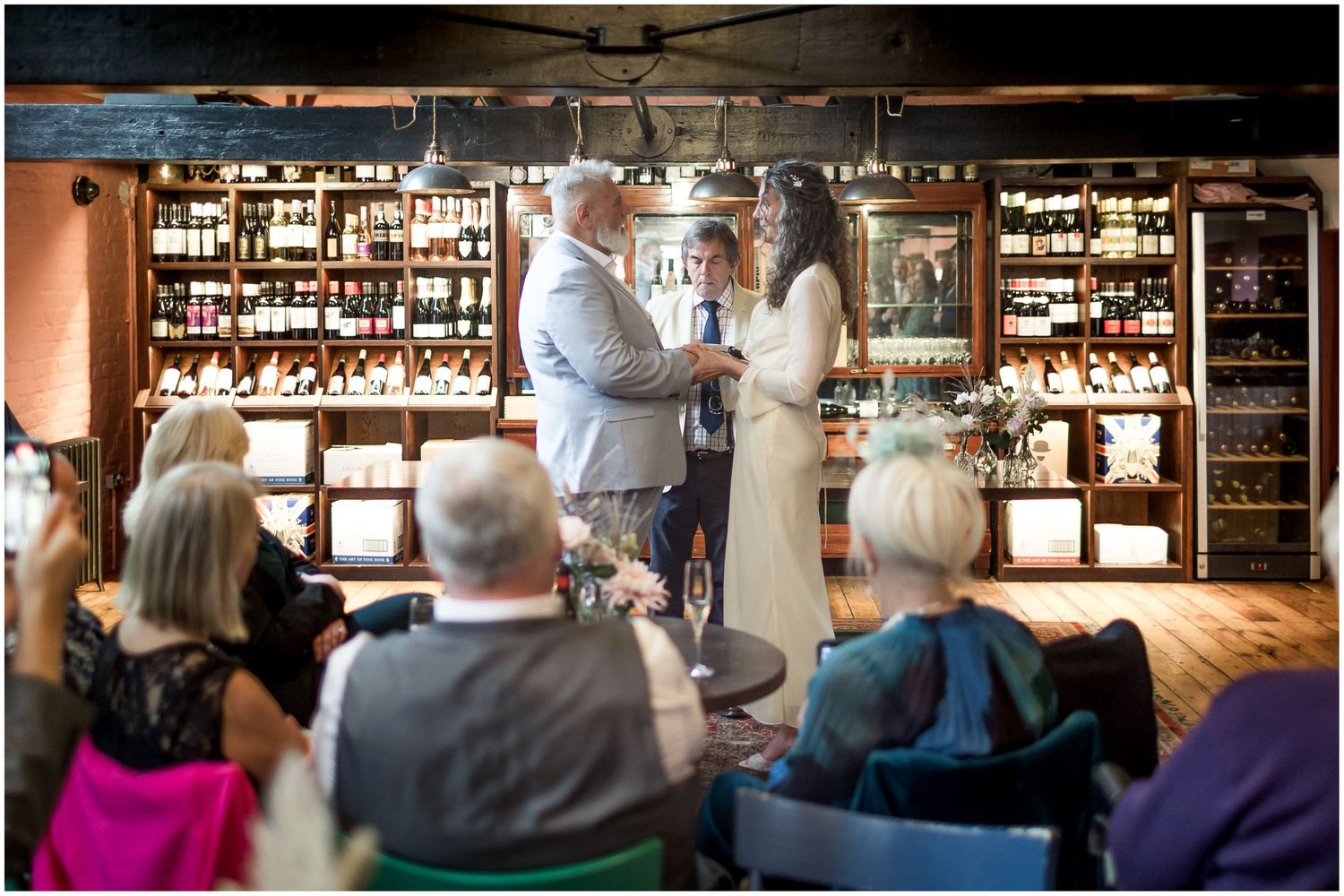 People gather at a dimly lit bar counter with shelves of bottles lining the wall behind them.