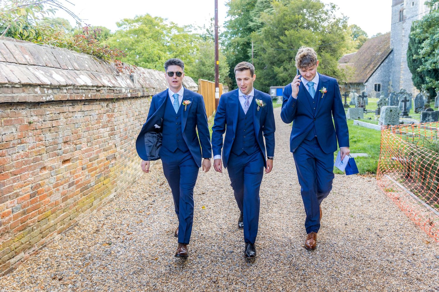 Three groomsmen in matching navy suits walk along a brick-walled path.