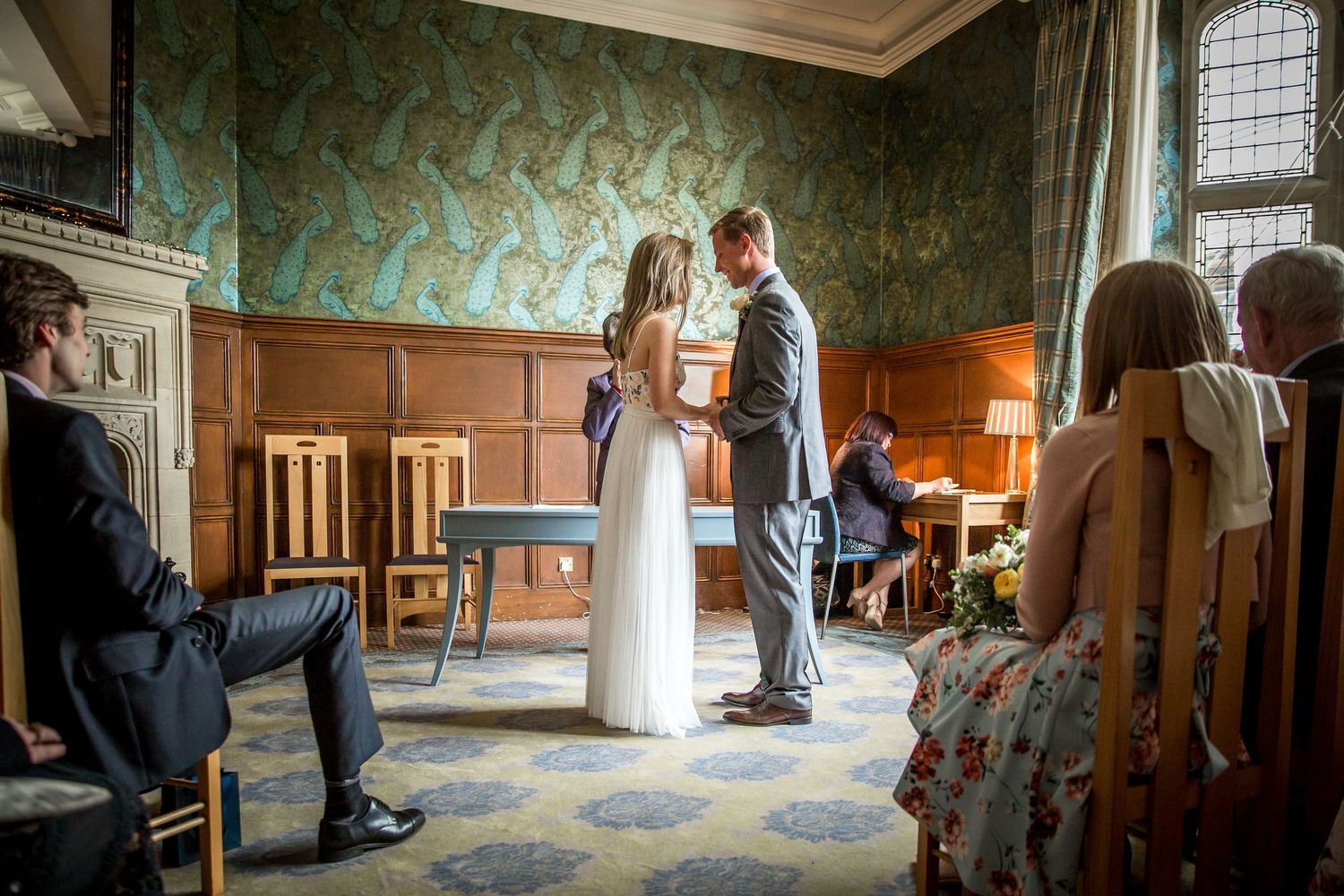Couple in formal attire stands in an ornate room with teal wallpaper and vintage furniture during a wedding ceremony.