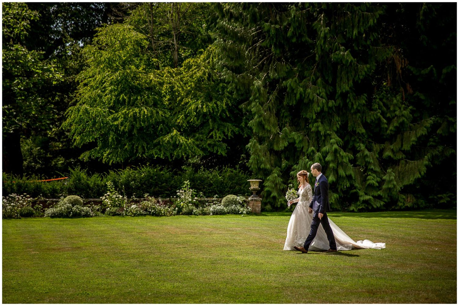A romantic couple shares a tender moment on a lush green lawn with trees in the background during a summer wedding.