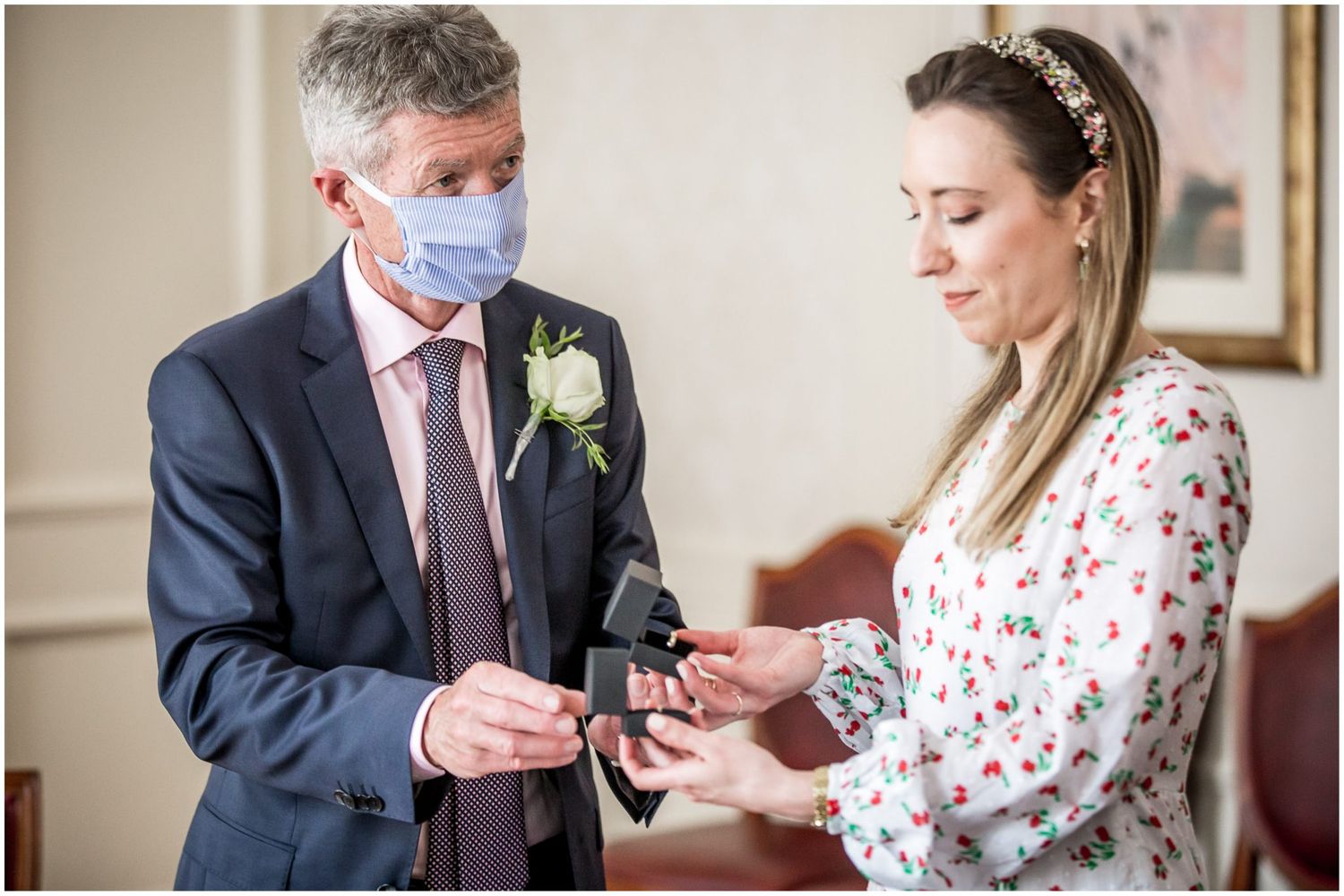 Close-up of hands exchanging rings during an indoor ceremony with floral dress details.