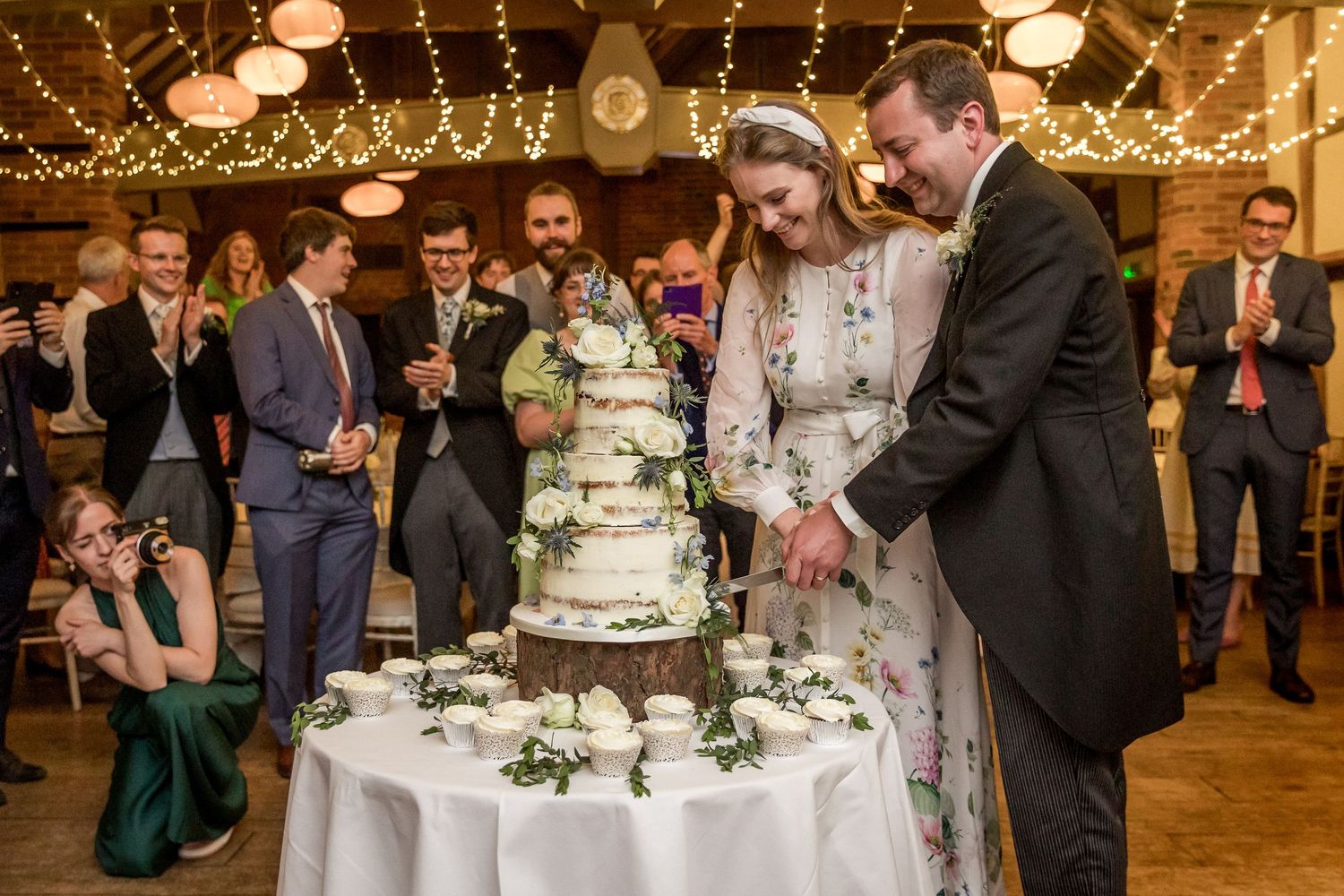 Wedding reception guests gather around an elegant tiered white cake decorated with flowers under warm lighting.