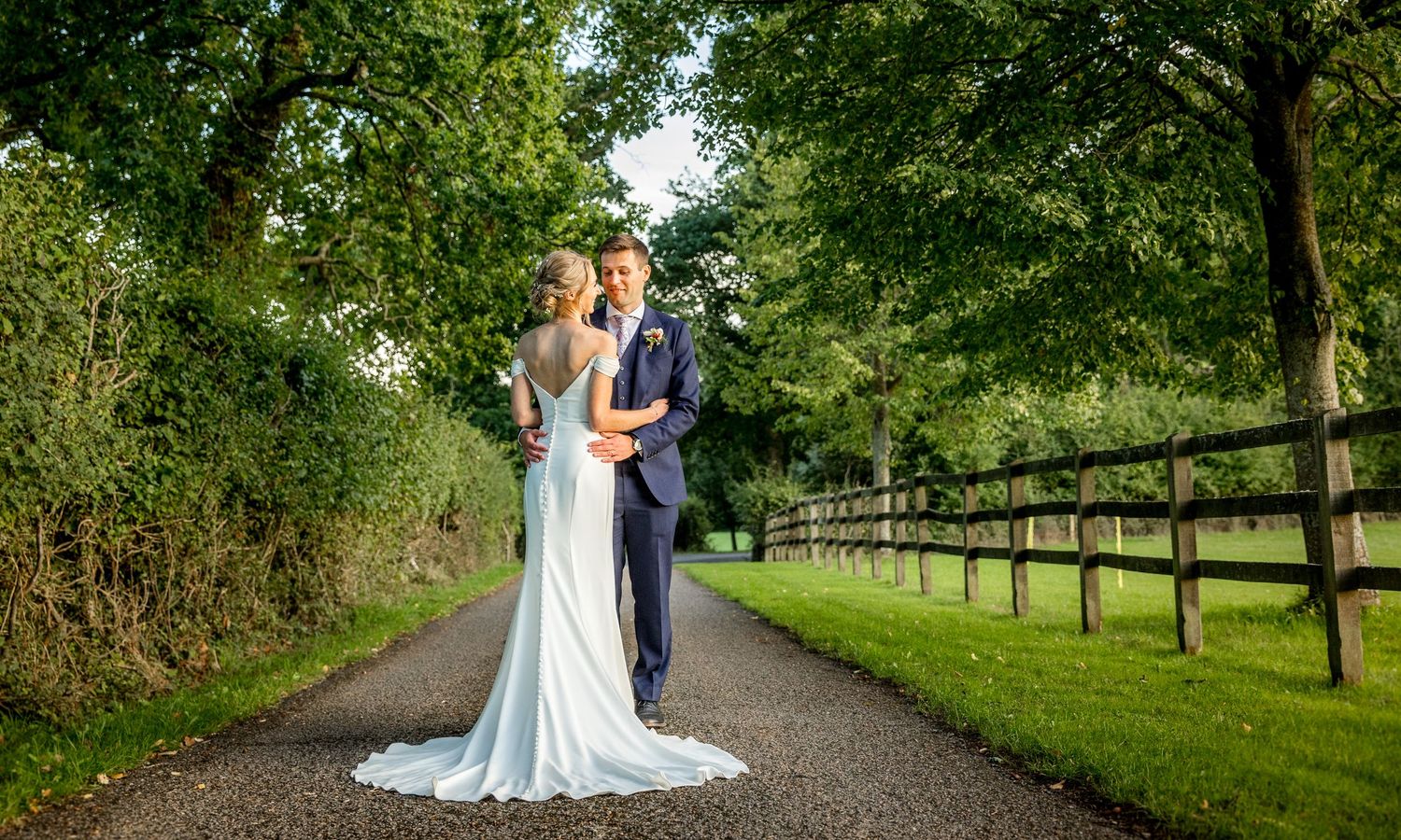 A romantic wedding couple walking along a tree-lined path in a lush garden setting.