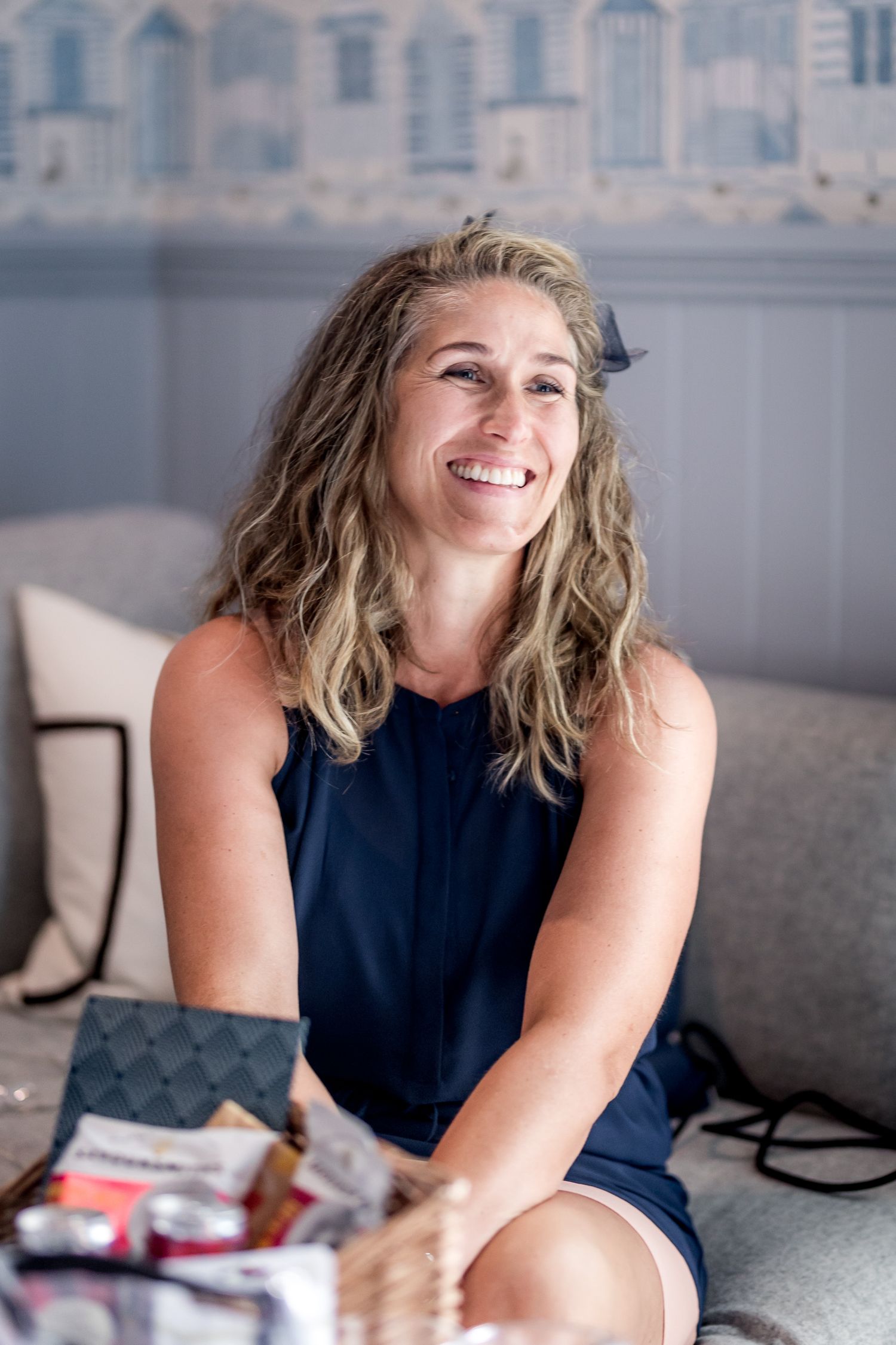 Woman in navy blue dress sitting casually on couch with bright smile.