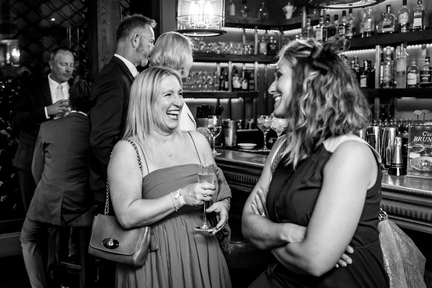 Black and white candid shot of guests laughing at bar during reception.
