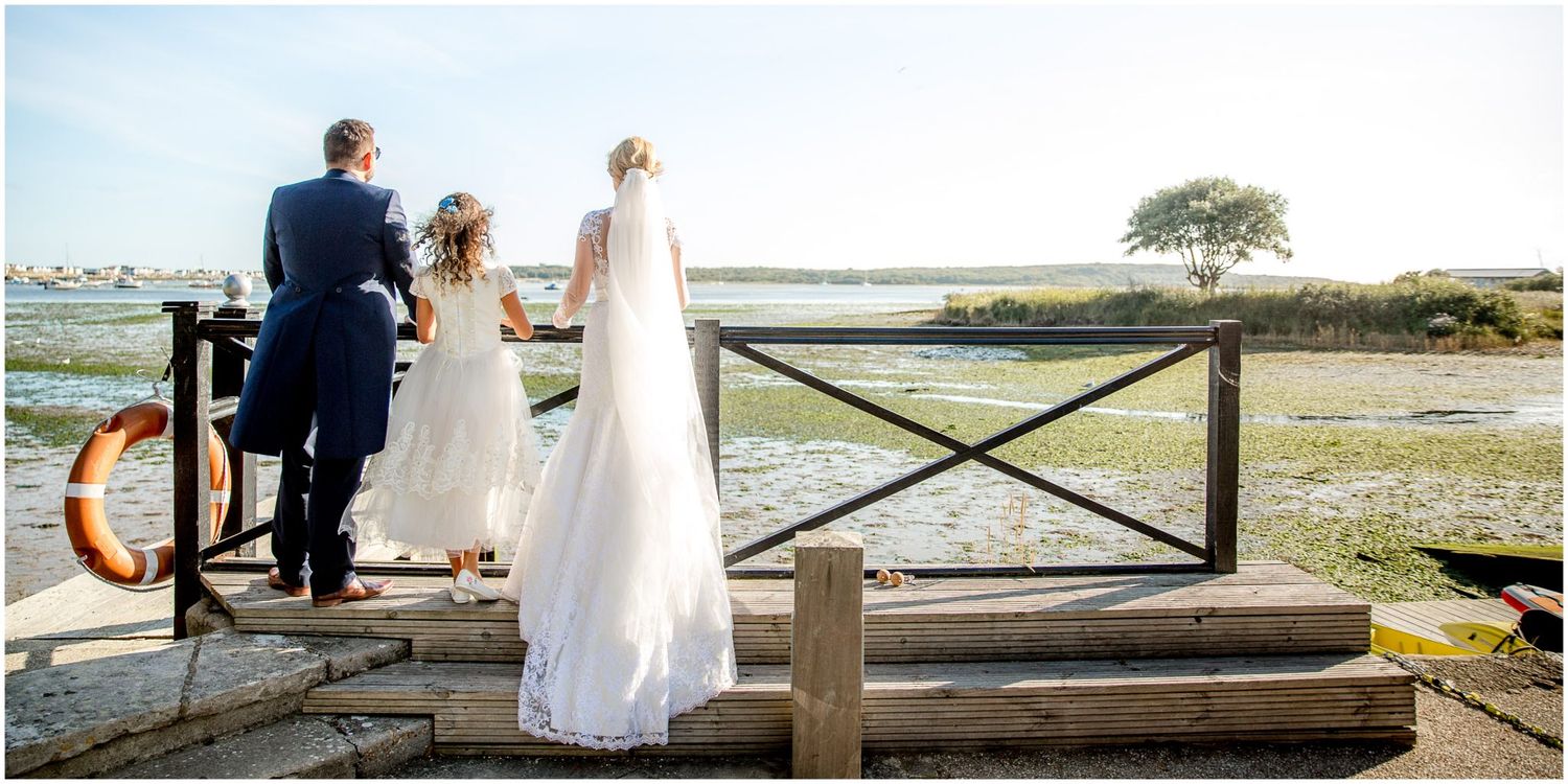 A couple in wedding attire stands at a wooden fence overlooking a scenic coastal landscape at sunset.