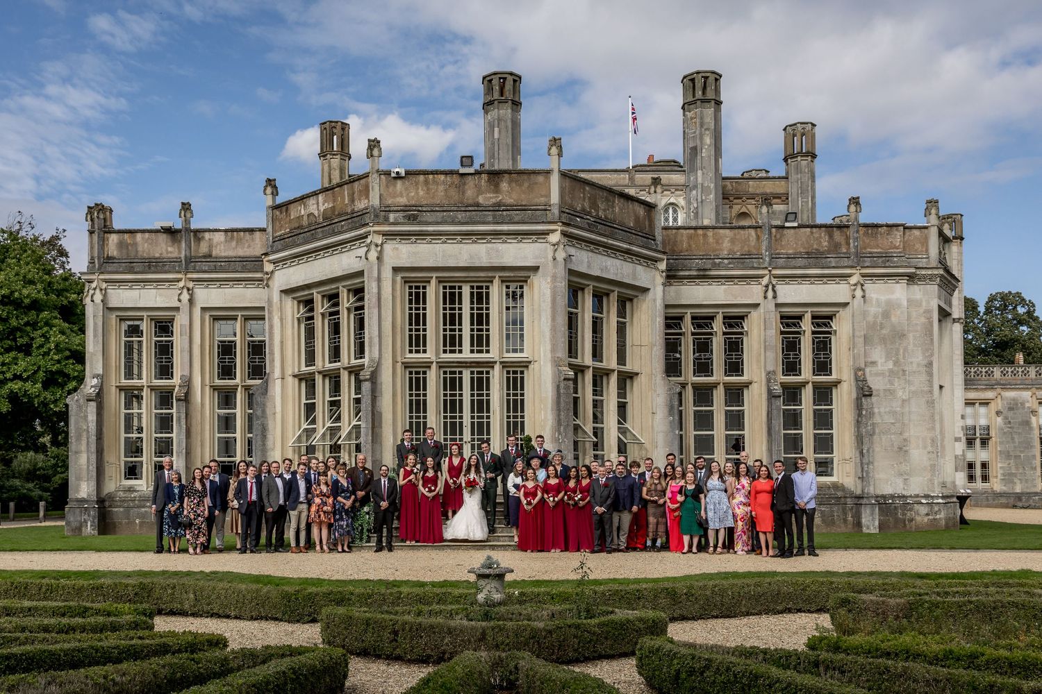 Wide shot of a historic English manor house with gathered guests on the front lawn.
