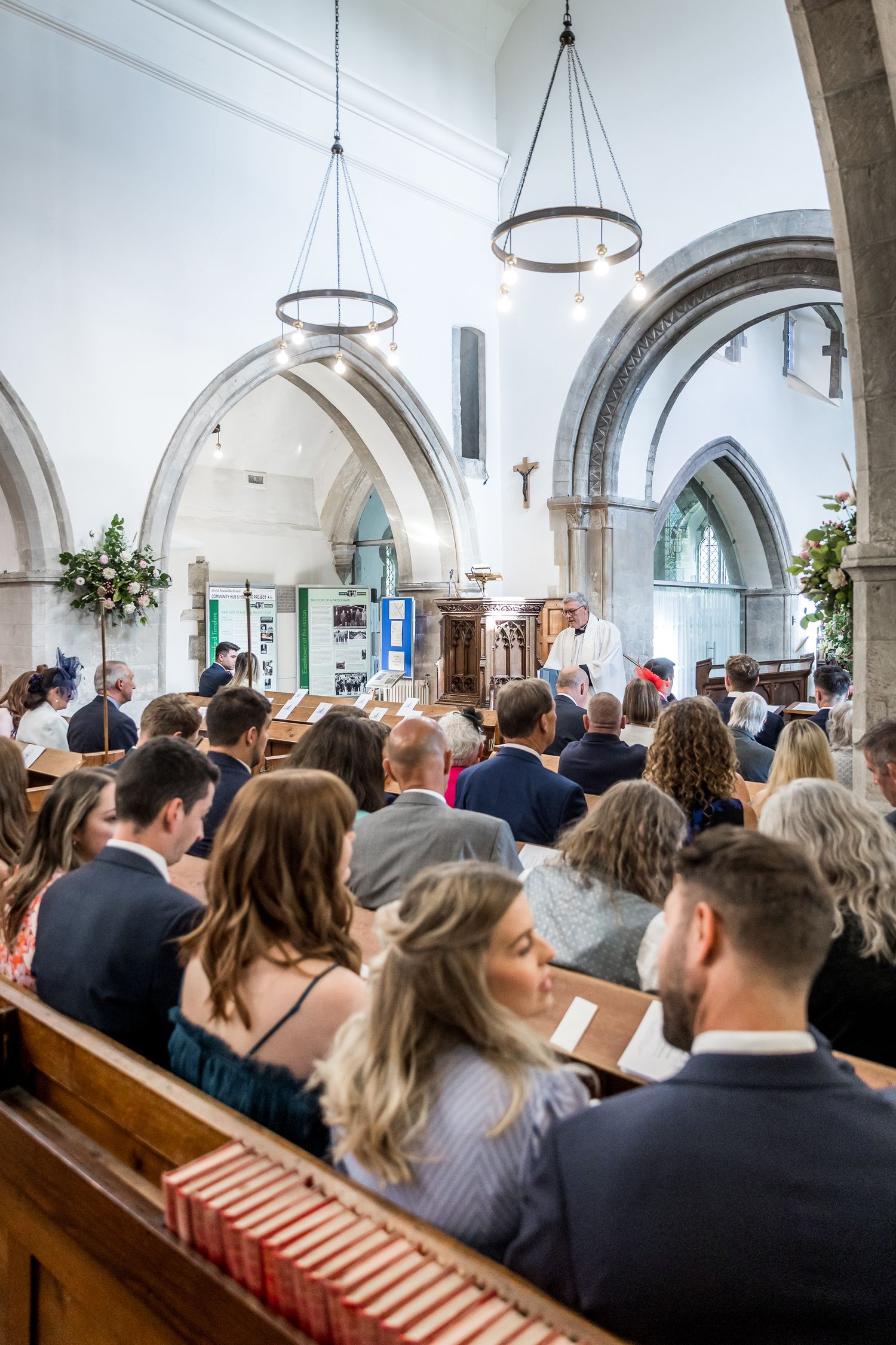 Wedding guests seated in church pews during ceremony under arched ceiling and chandeliers.