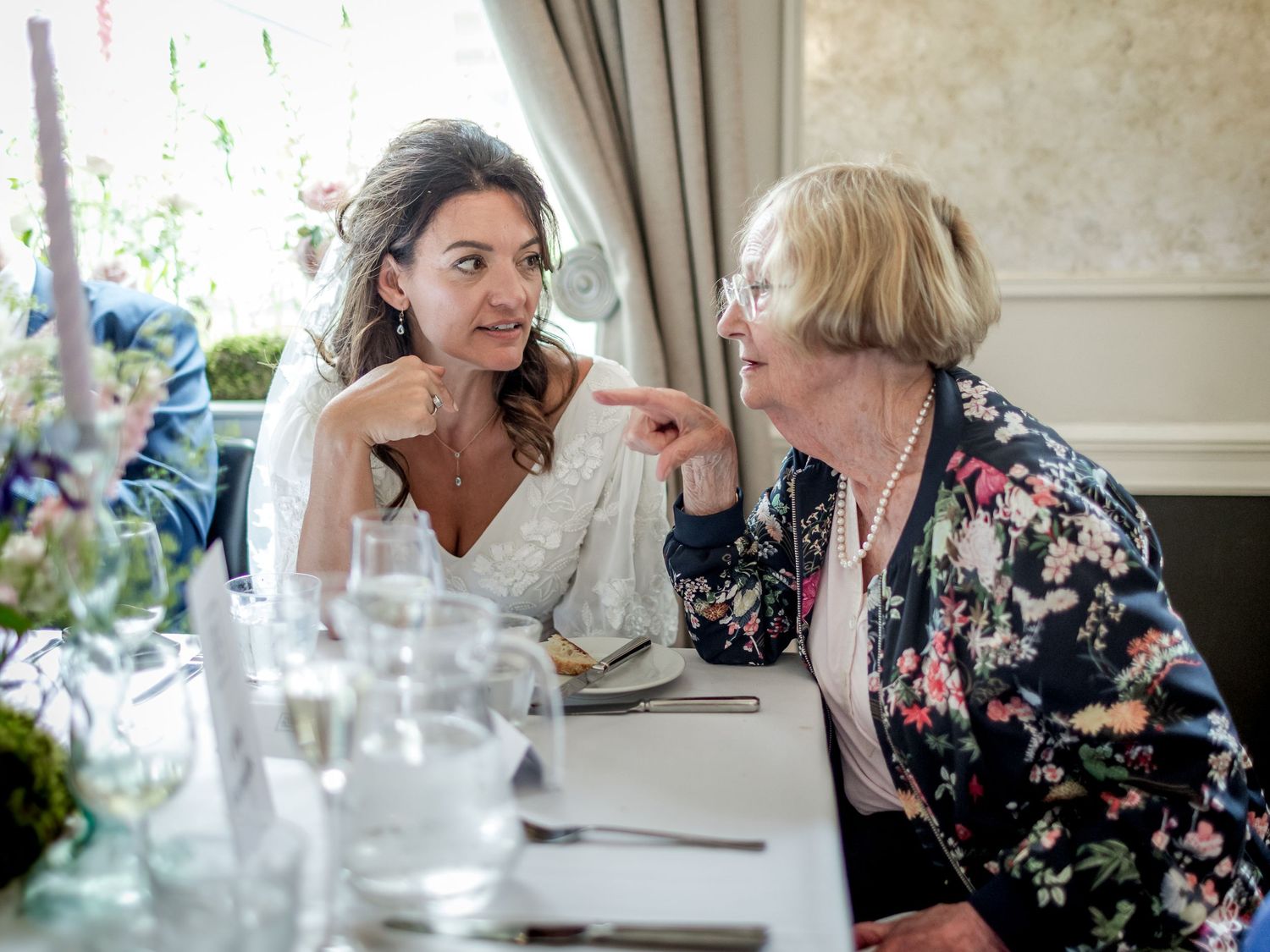 Wedding guests in floral dresses enjoy conversation during an indoor reception.
