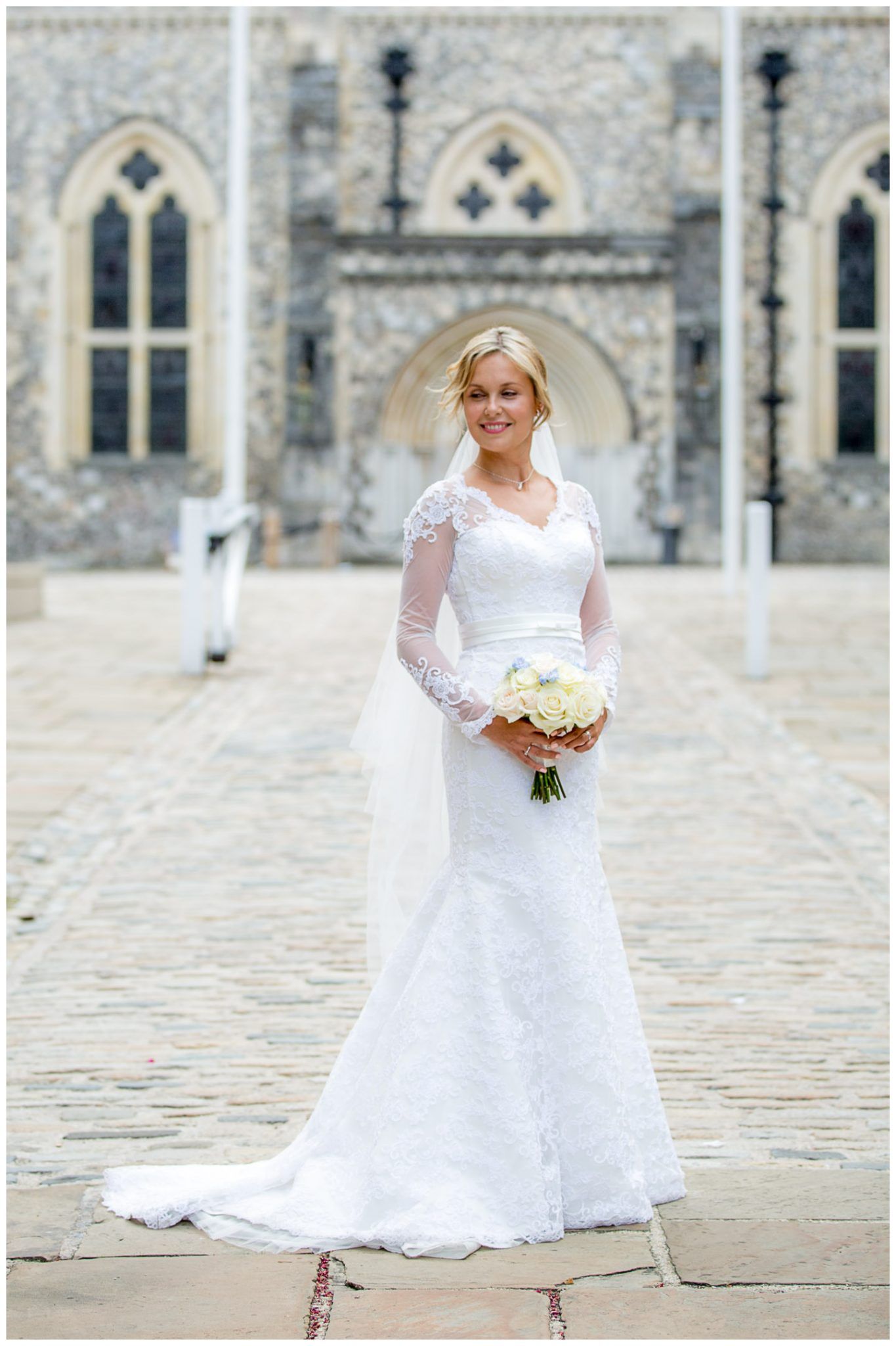 A bride in a white lace gown with long sleeves poses on cobblestone steps in front of a historic stone church.