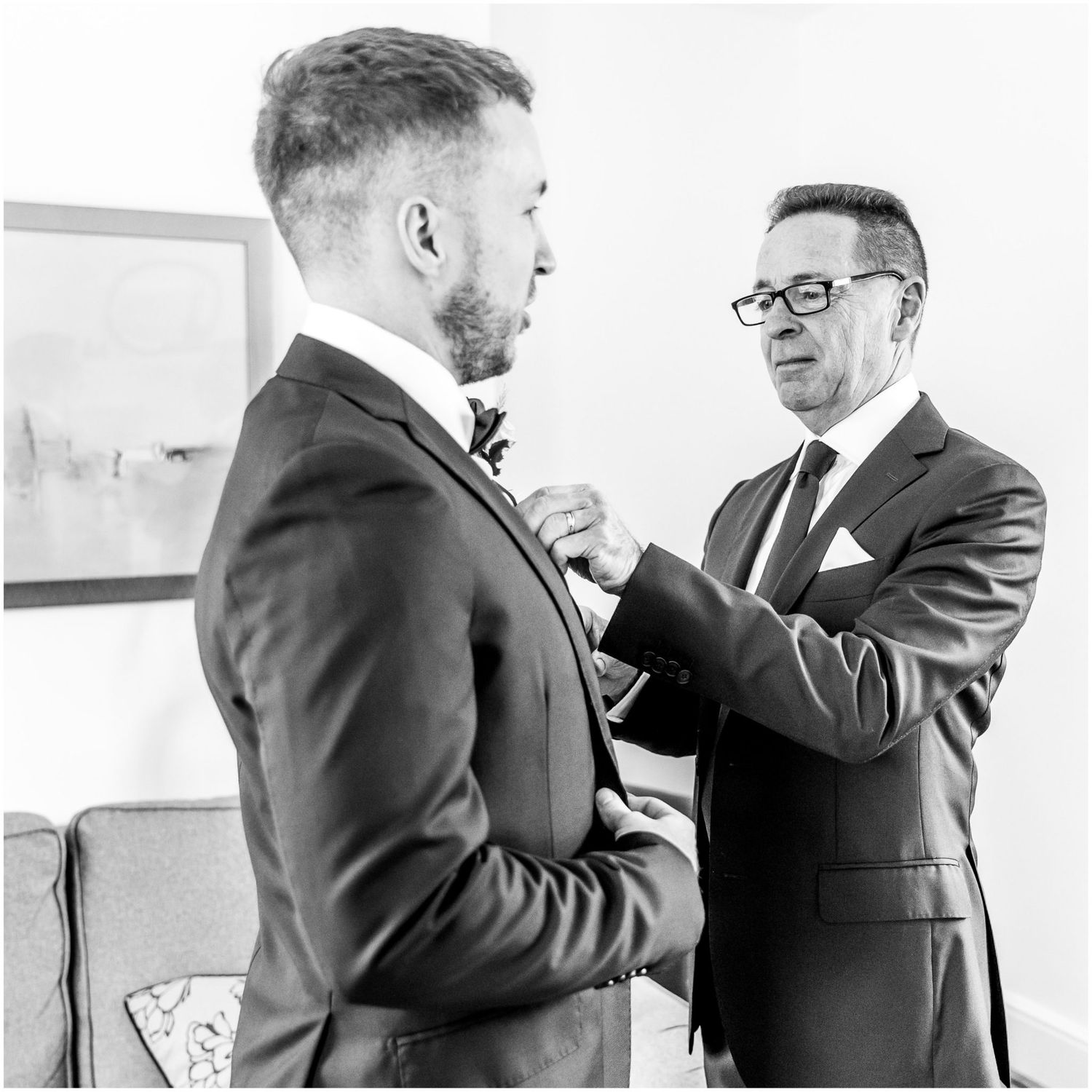 Two people in dark suits helping each other with formal attire preparations indoors.