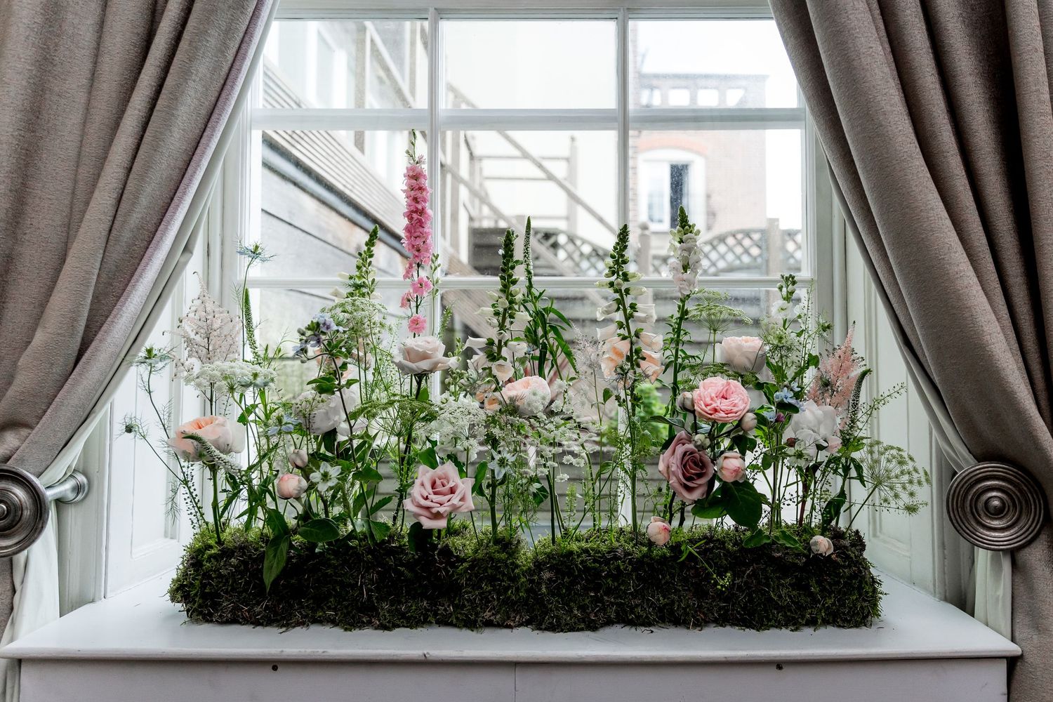 Window display with lush greenery and flowers against grey curtains.