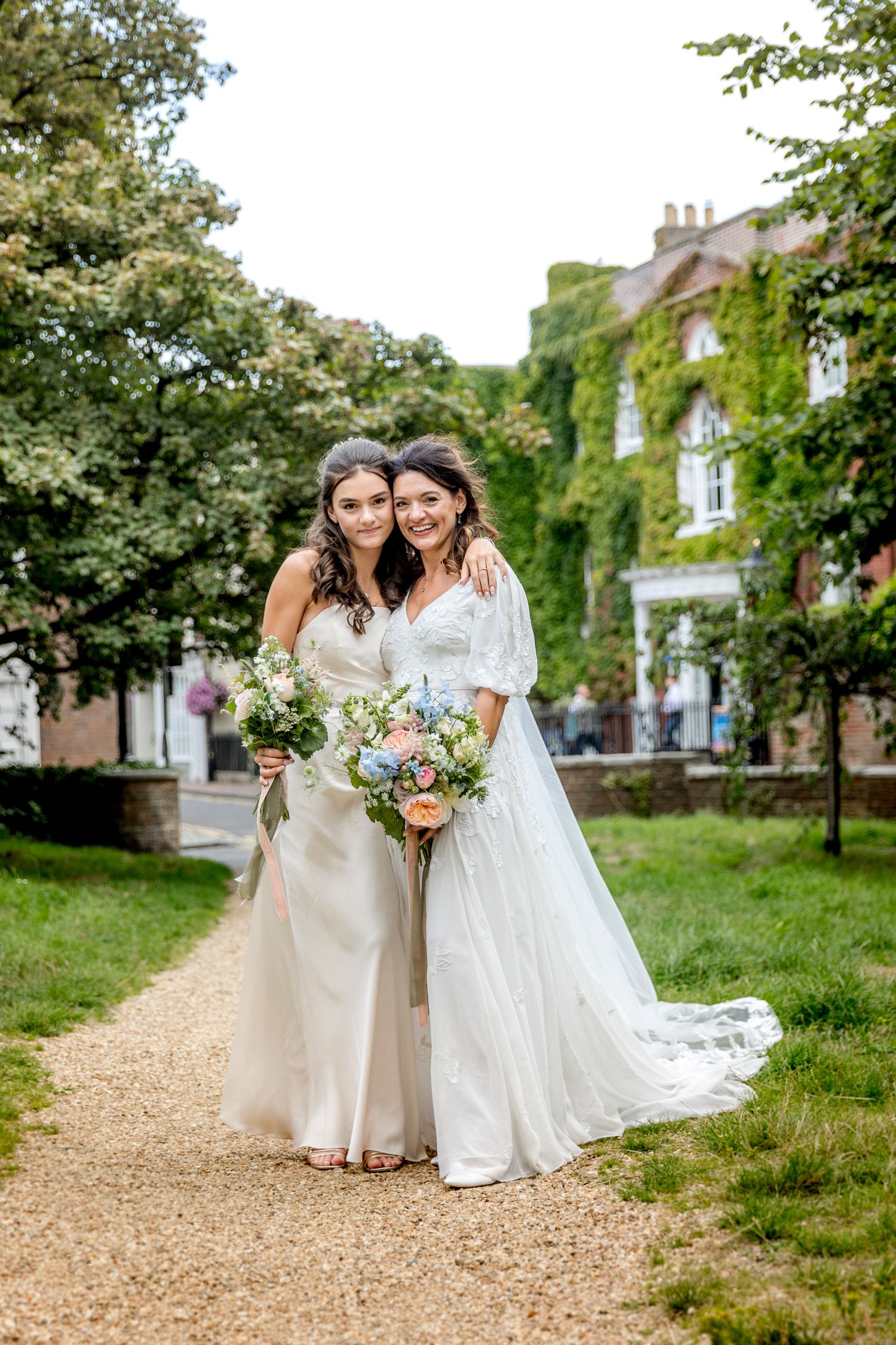 Two people in flowing white dresses share joyful moment on garden path.