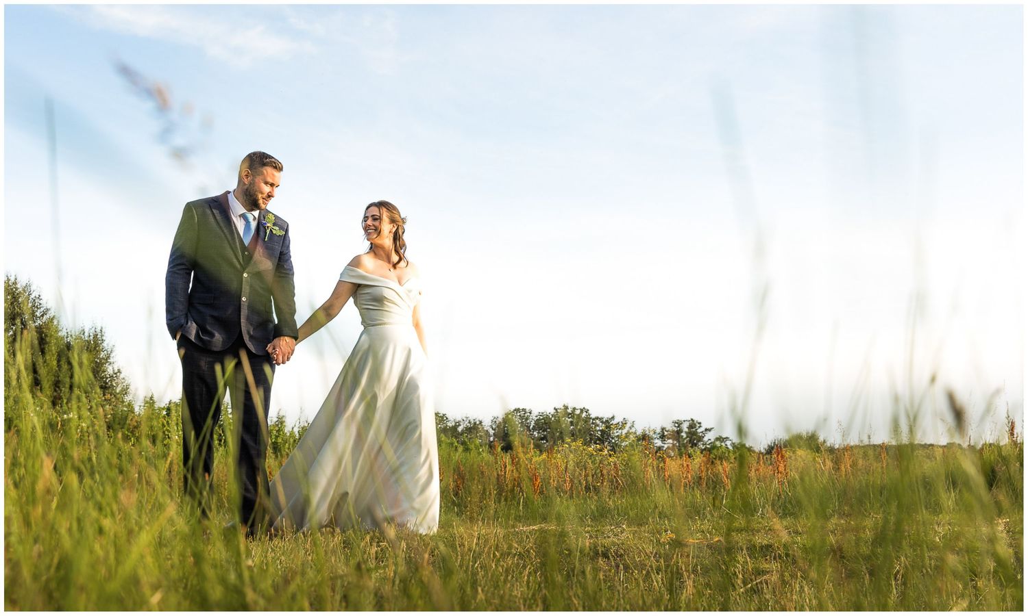 A bride and groom hold hands walking through a lush green meadow on their wedding day.