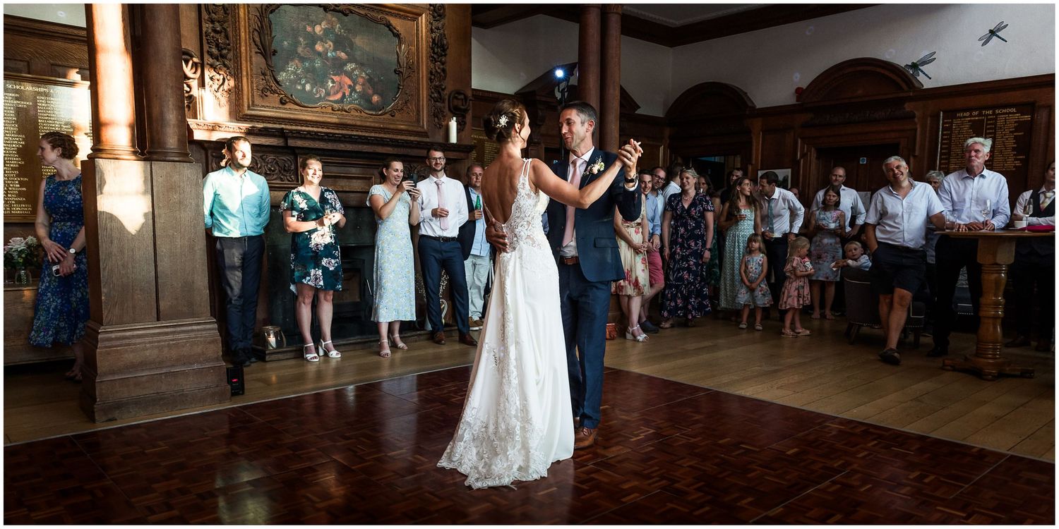Bride and groom share their first dance on a wooden floor surrounded by wedding guests at Walhampton School