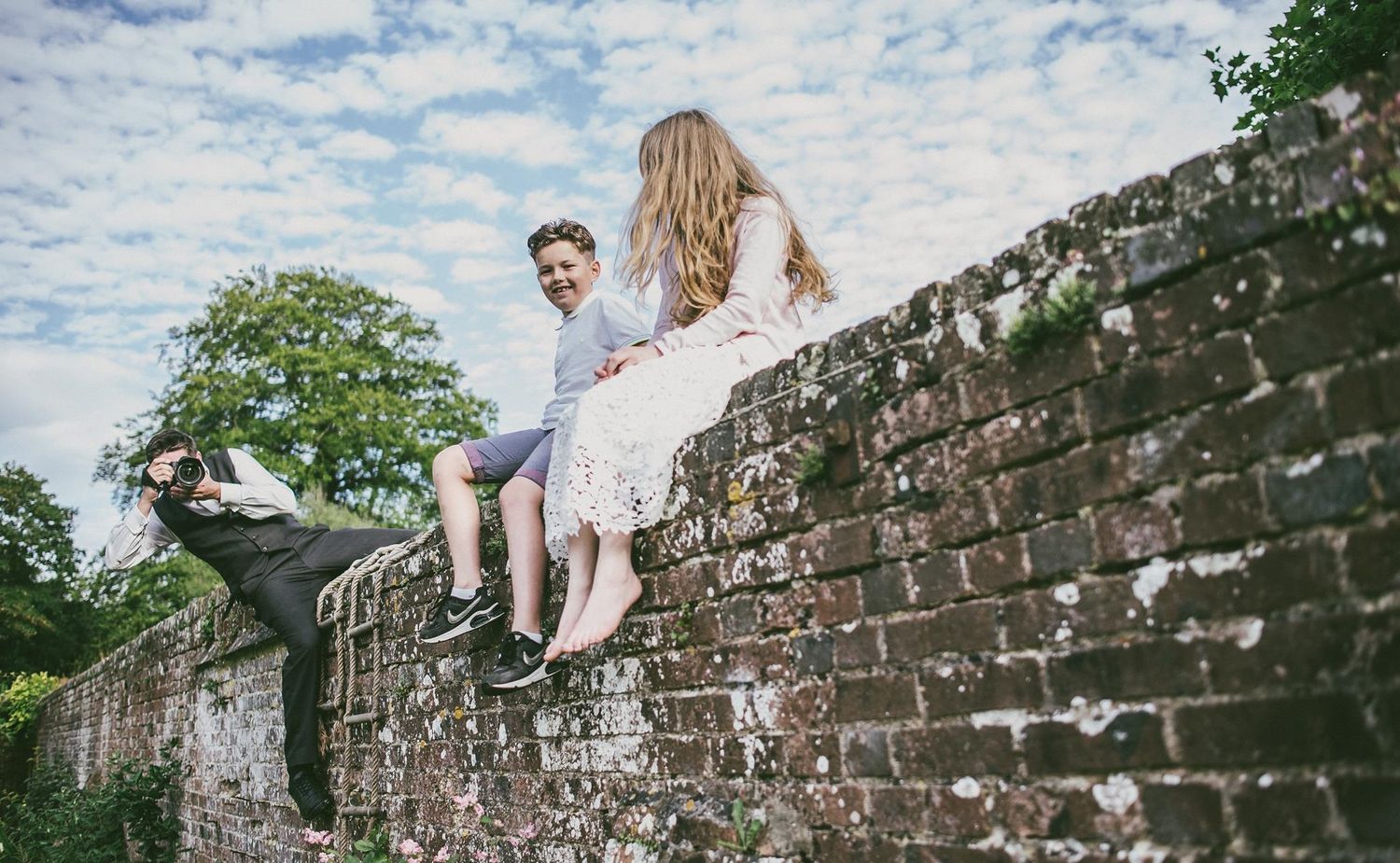 Wedding couple sits atop rustic brick wall under blue sky with wispy clouds.
