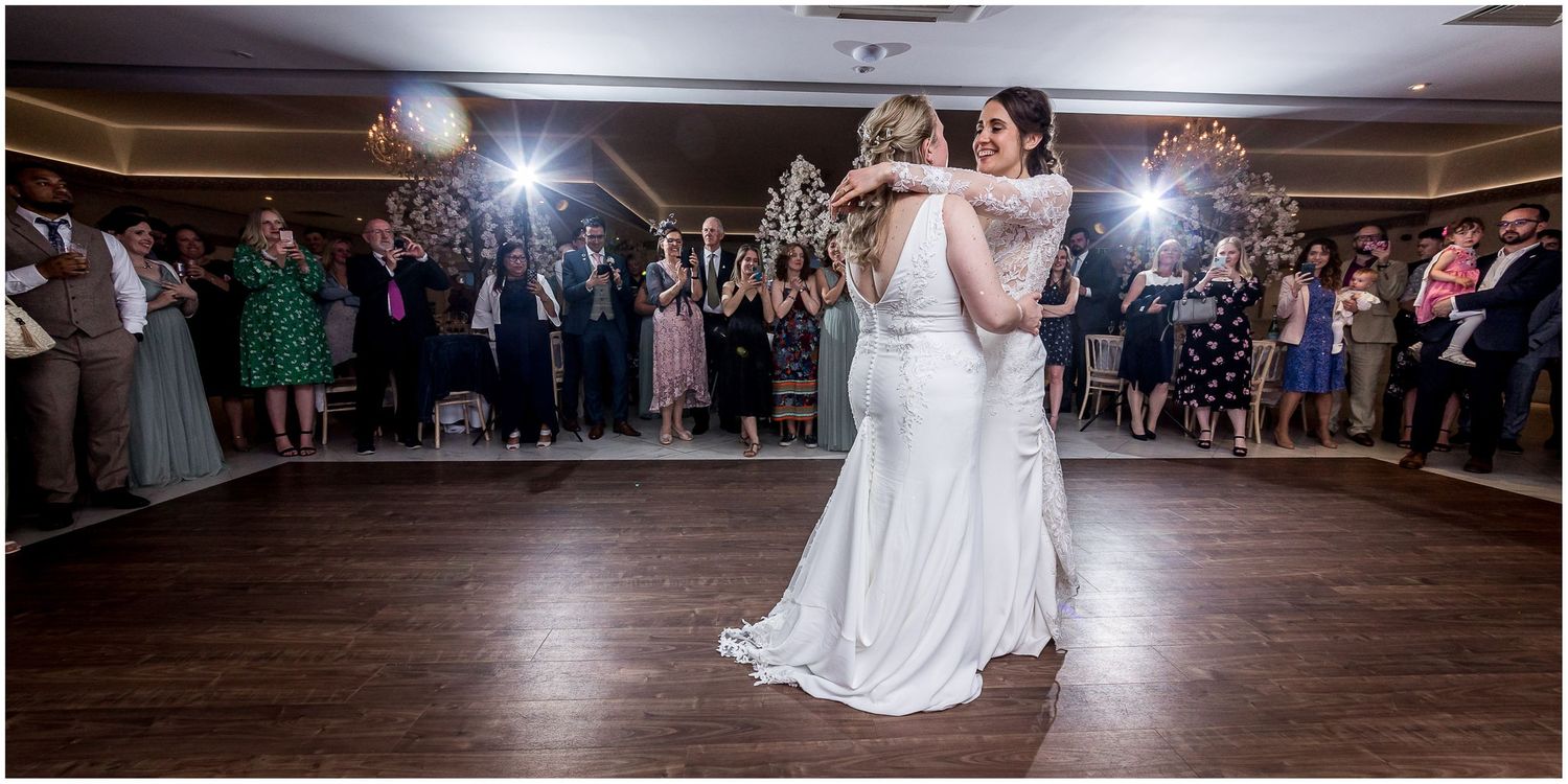 Couple sharing first dance on wooden ballroom floor surrounded by guests.