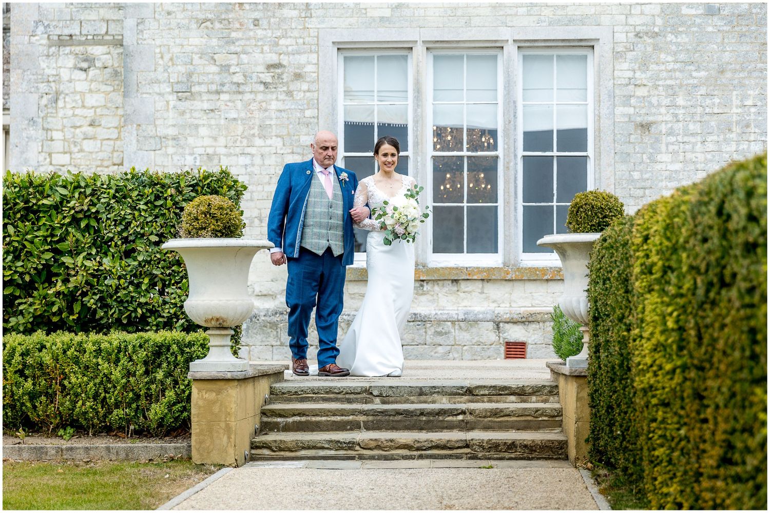 Wedding couple and guests pose in front of a classic white brick mansion with manicured gardens.