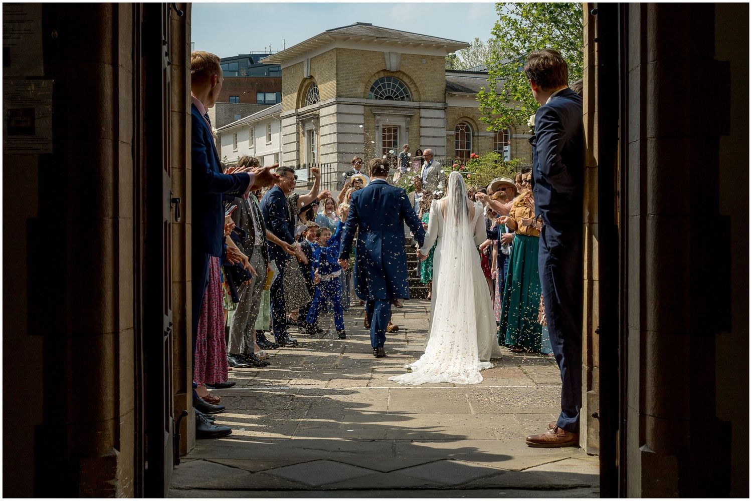Wedding party and guests gathered outside historic stone church entrance on sunny day.