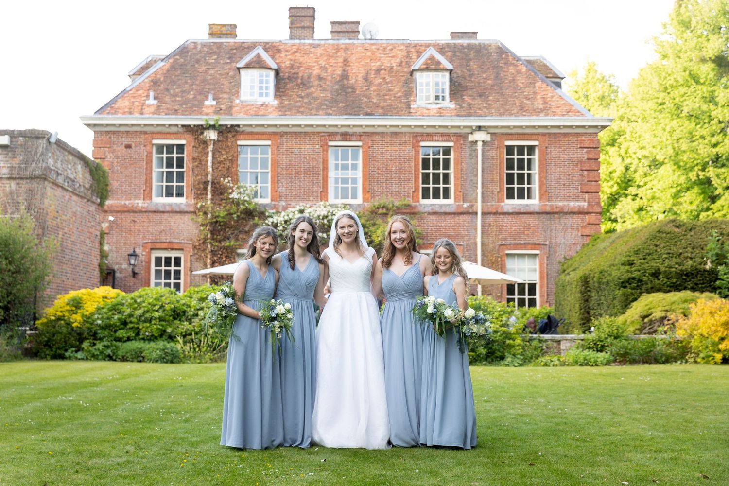 Wedding party in light blue dresses poses on the lawn of an elegant brick estate.