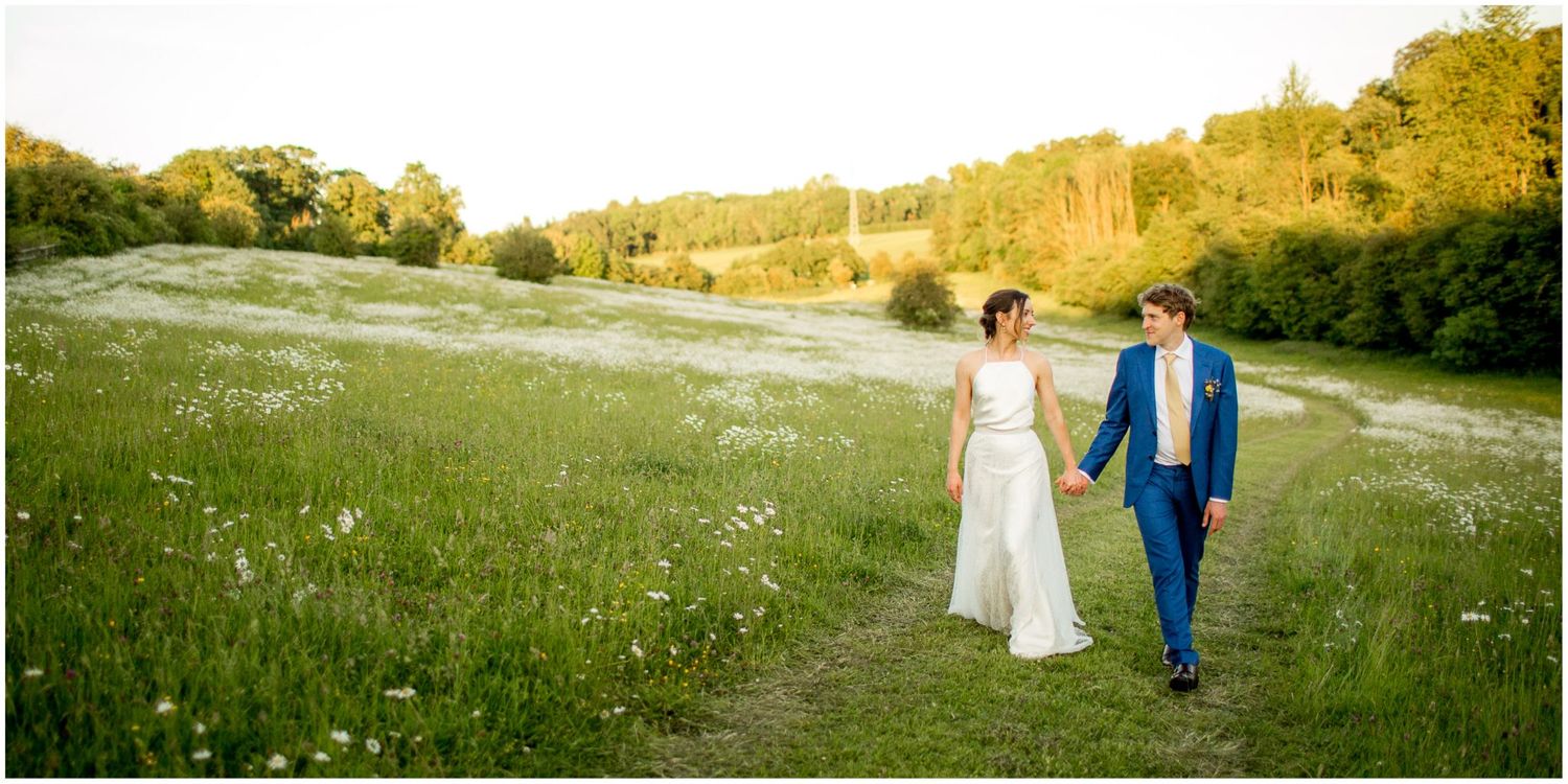 Newlyweds walk hand in hand through a serene meadow with golden evening light.