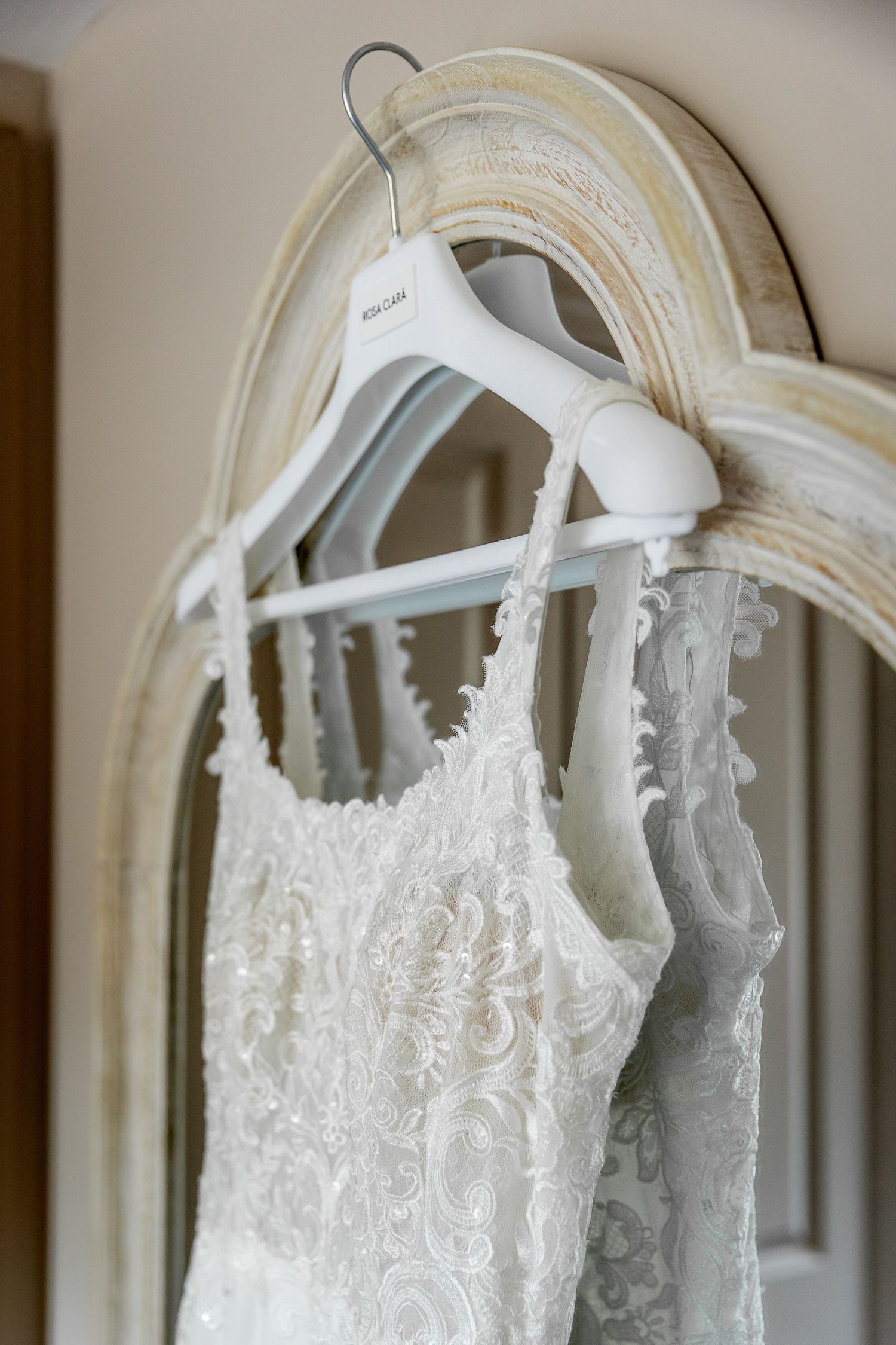 White lace wedding dress hanging on ornate mirror with delicate detail visible.